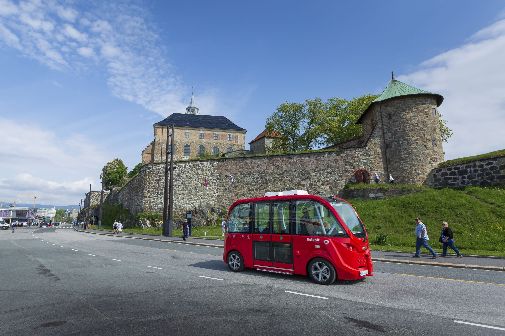 Red autonomous shuttle bus on road near historic stone fortification, with people walking and partly cloudy sky.