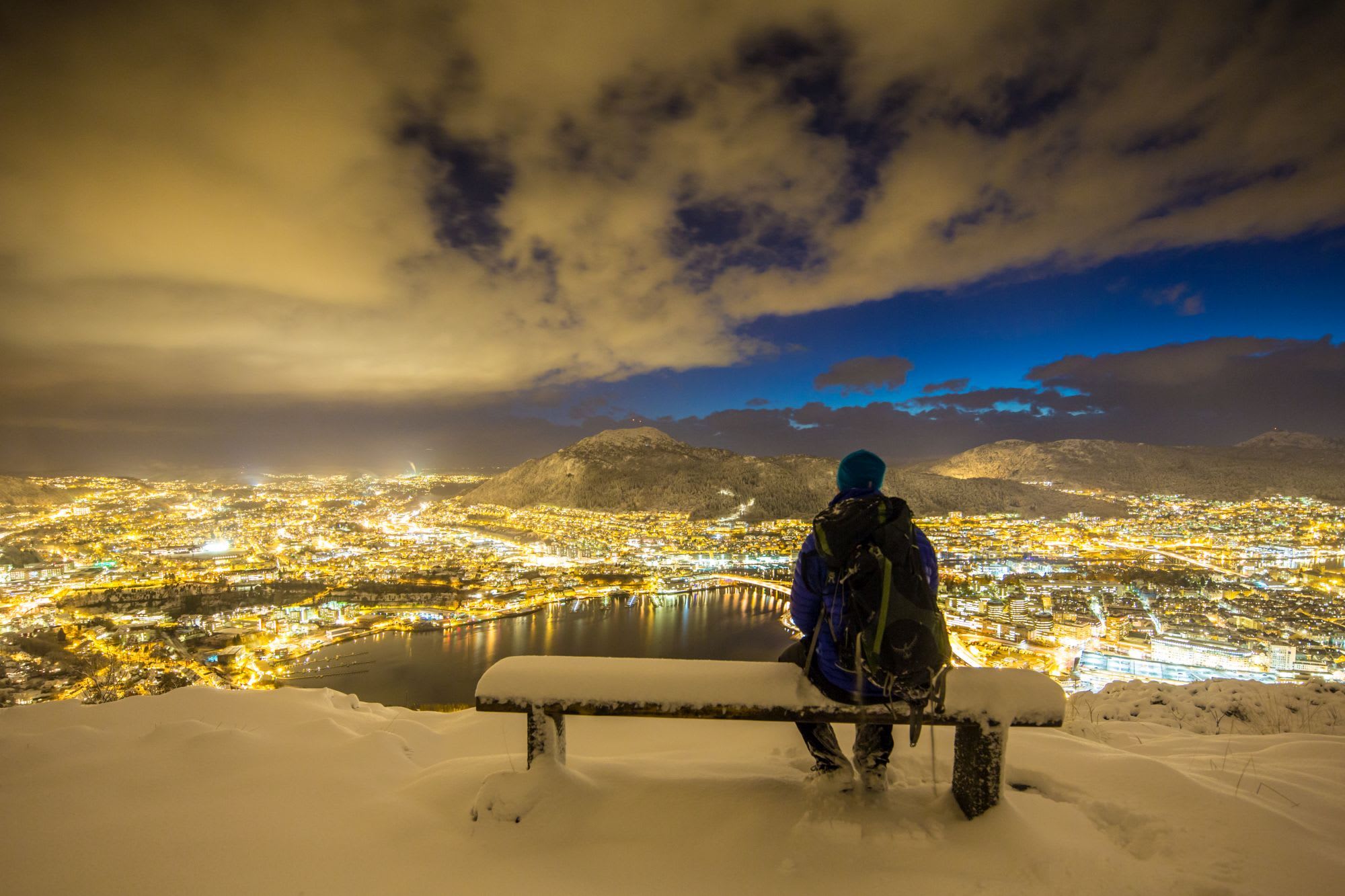 Person sitting on snow-covered bench overlooking Bergen’s city lights at night, with snowy mountains and soft blue sky.