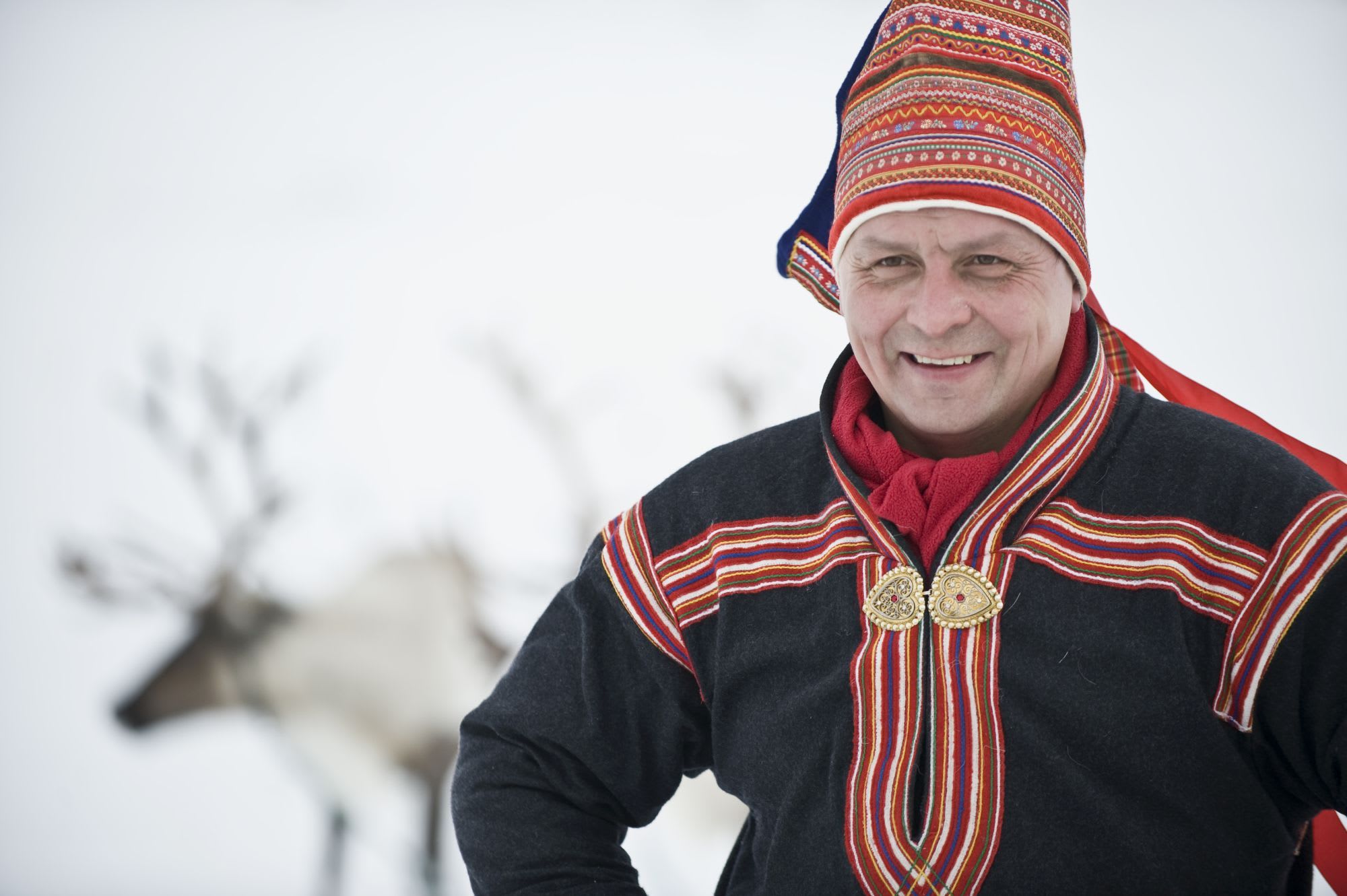 A smiling man in traditional colorful clothing with intricate embroidery and a tall, pointed hat with red and gold details.