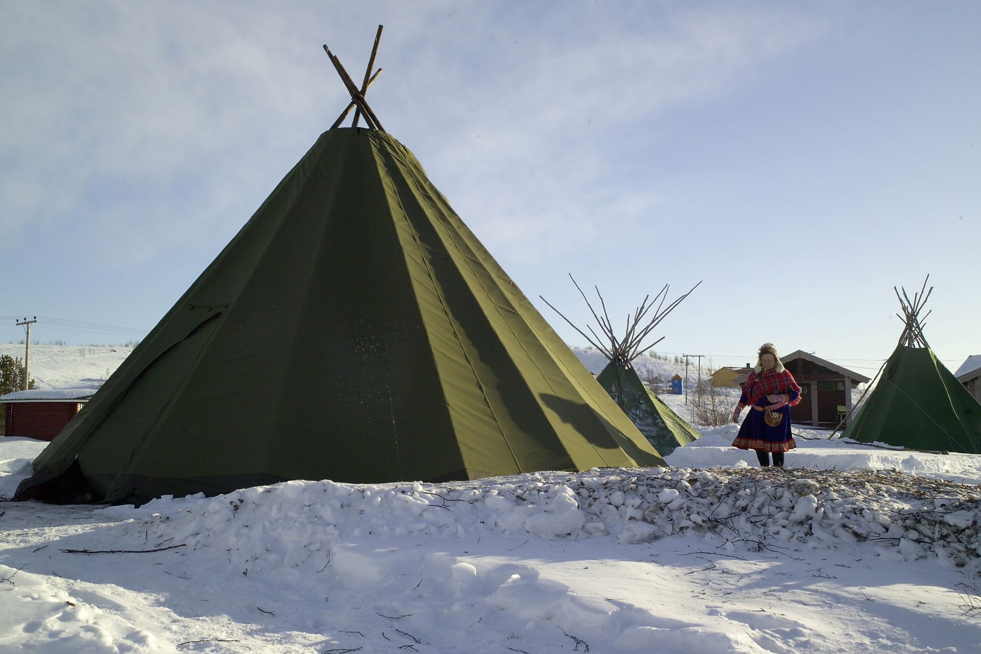 Person in Sami clothing standing next to "lavvu" tents in snowy landscape, clear blue sky, and serene, remote feeling.
