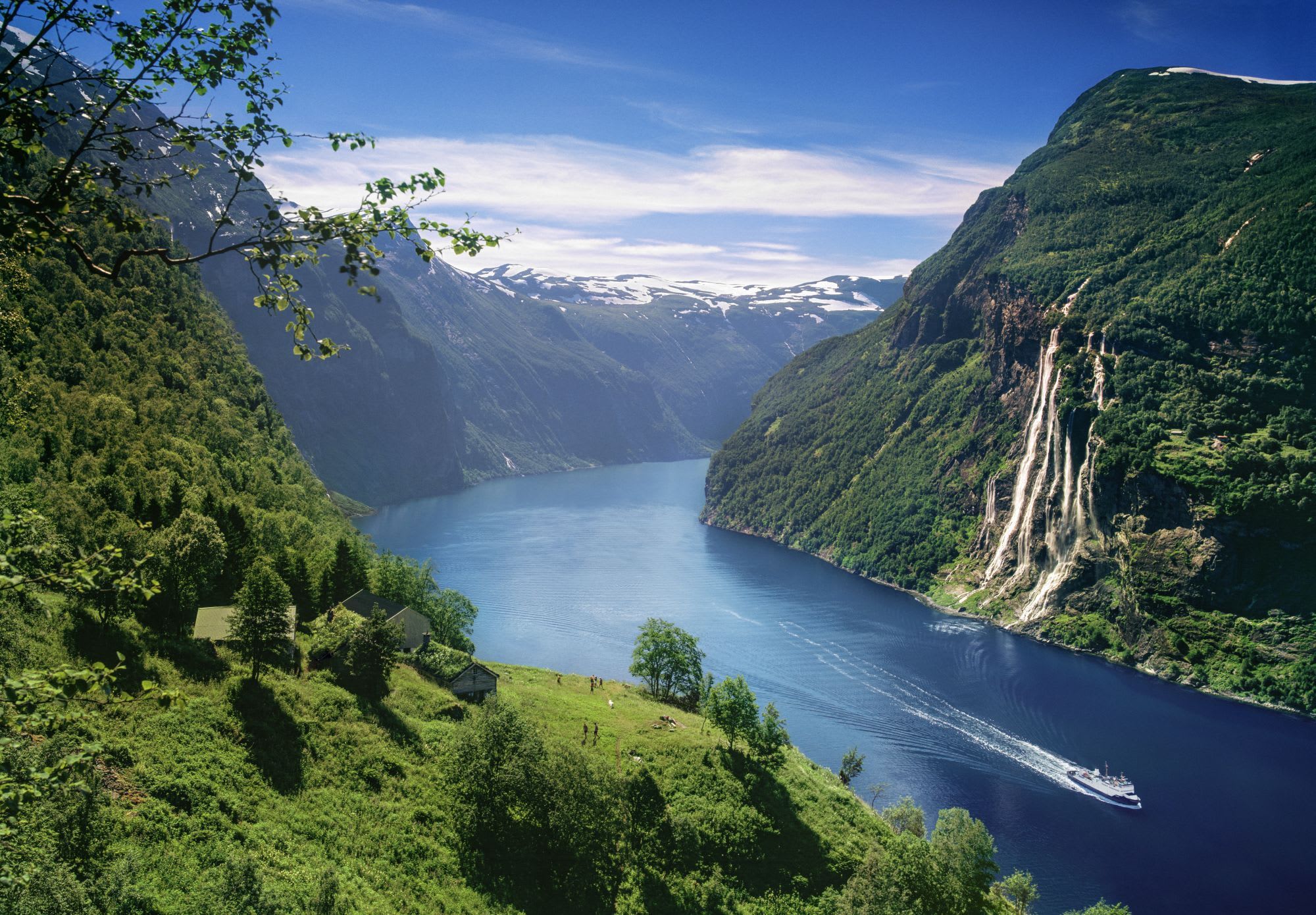 Norwegian fjord with steep cliffs, waterfalls, a boat cruising, and snow-capped mountains, reflecting in calm waters.