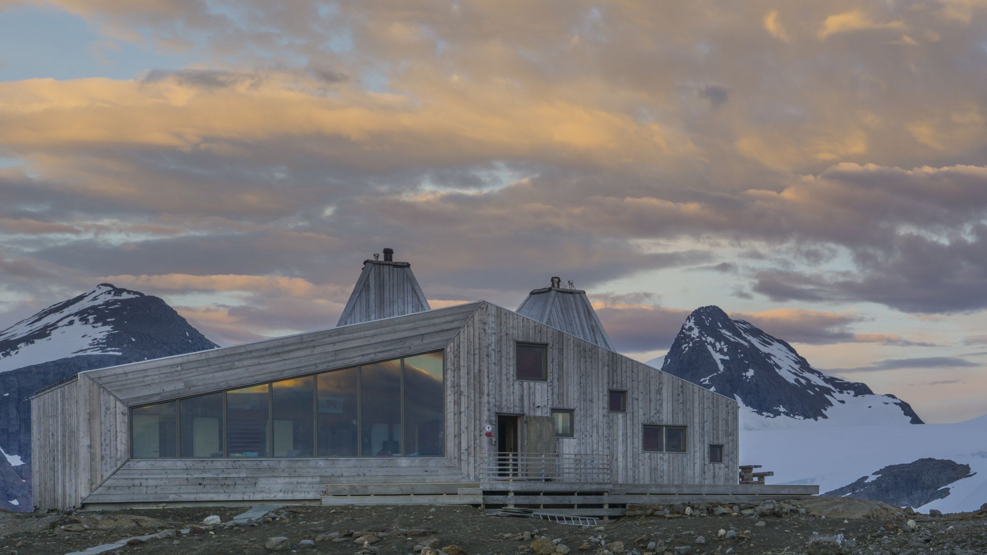 Modern angular wooden building with large windows, set against snowy mountains and pastel sunset sky.