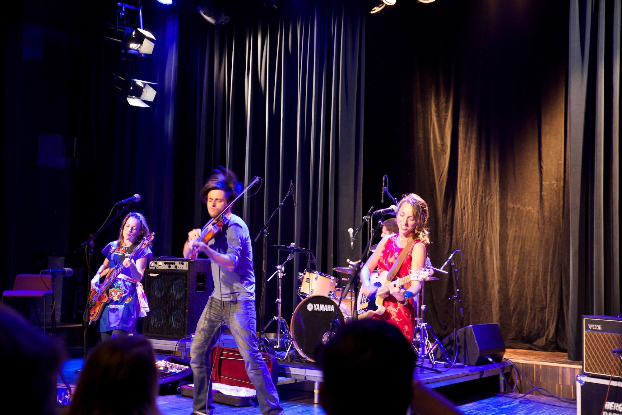Live music performance with violin, guitar, and bass players, warmly lit venue and dark backdrop with audience hints.