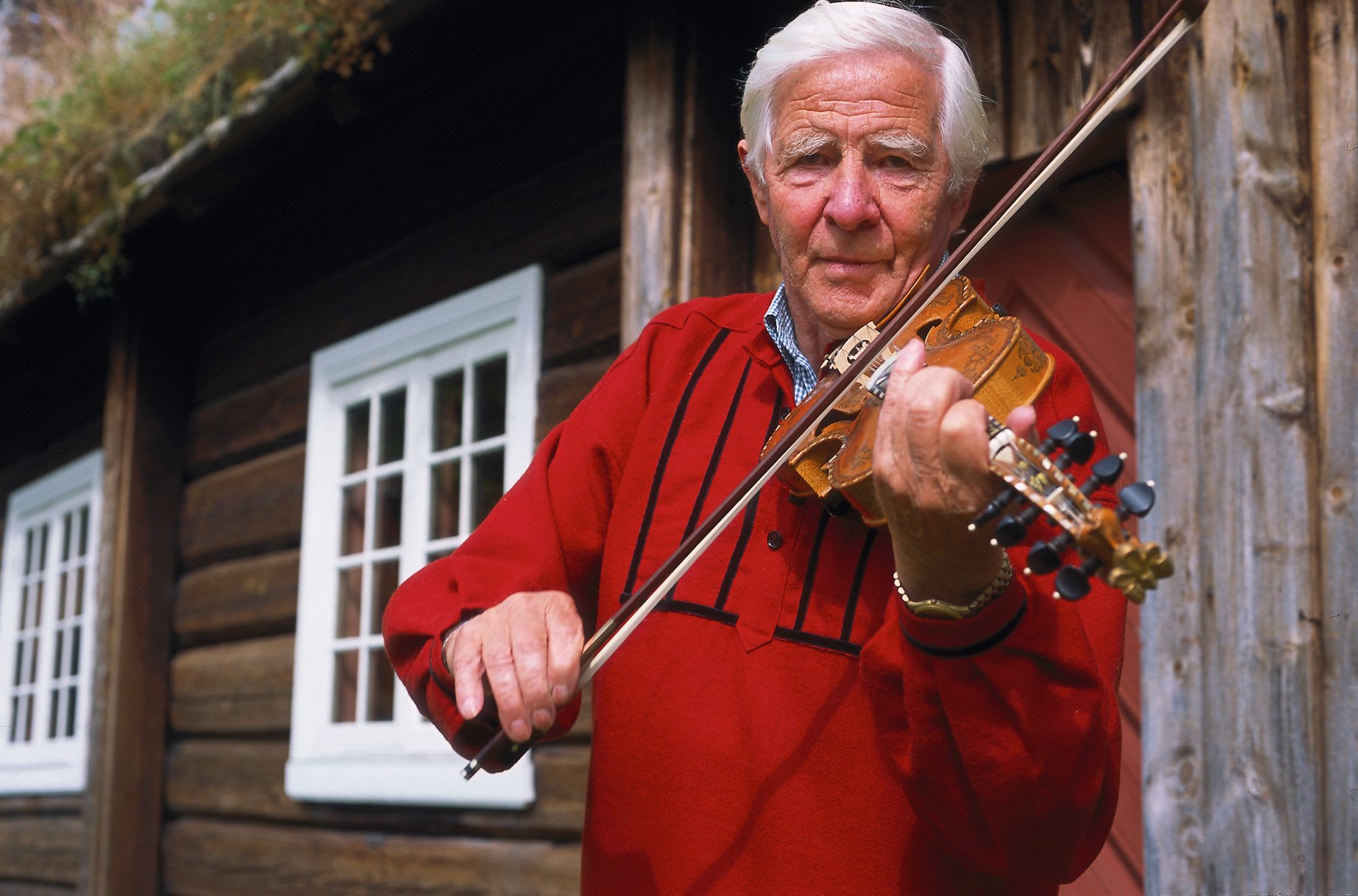 An elderly man plays a Hardanger fiddle in a red folk costume with black trim in front of a rustic building.