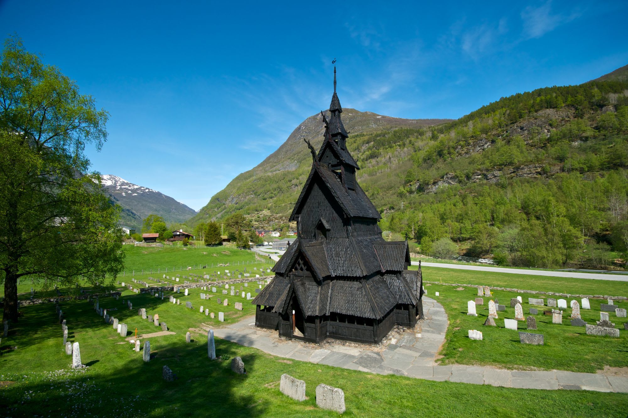 Preserved wooden stave church with dark carvings, surrounded by green landscape, gravestones, hills, trees, and snow-capped mountains.