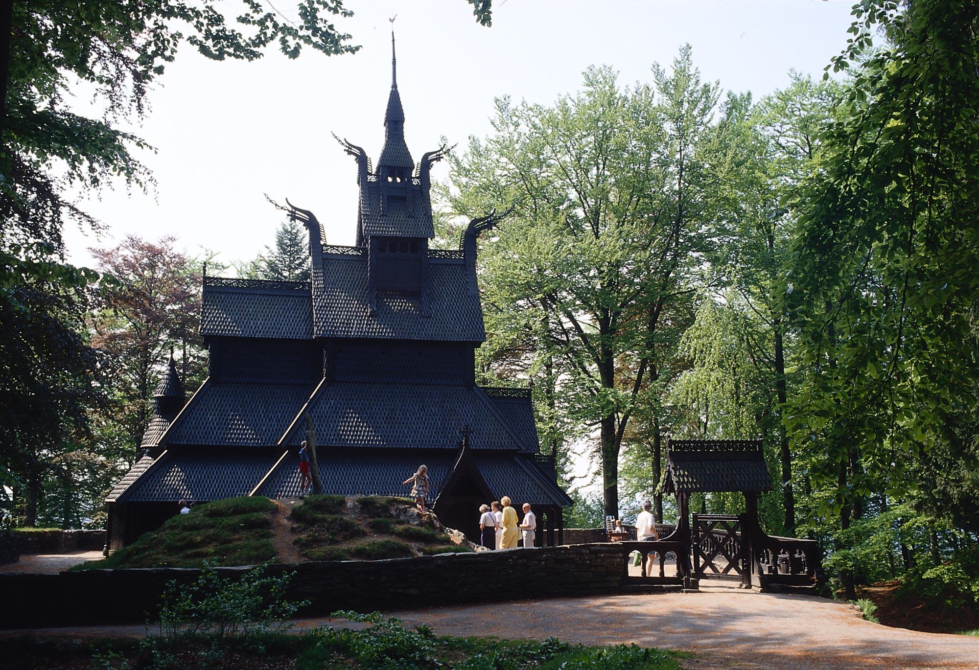 A black stave church surrounded by trees, with intricate carvings and a multi-tiered roof. Children play nearby.