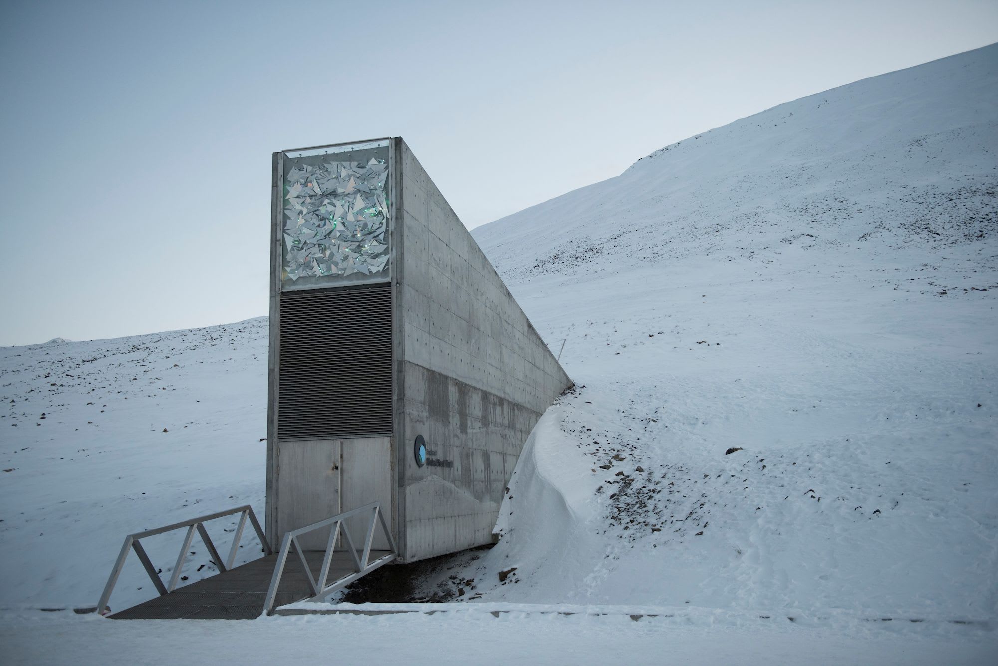 Minimalist concrete structure partially buried in snowy hillside, with decorative glass artwork at entrance under pale sky.