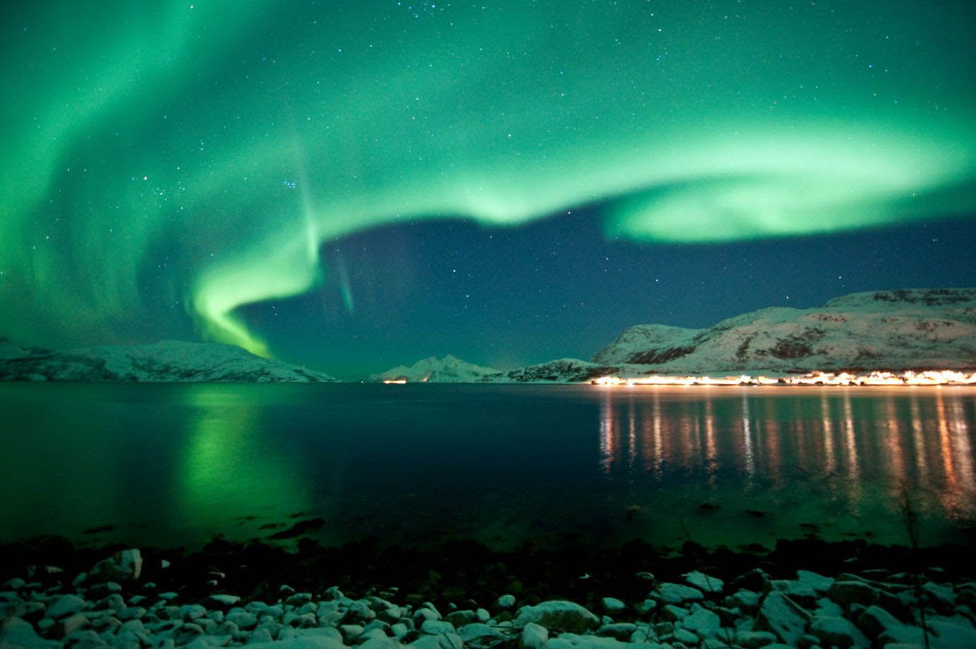Northern Lights in vivid green hues swirling over dark fjord with snow-capped mountains, village lights reflected on water.