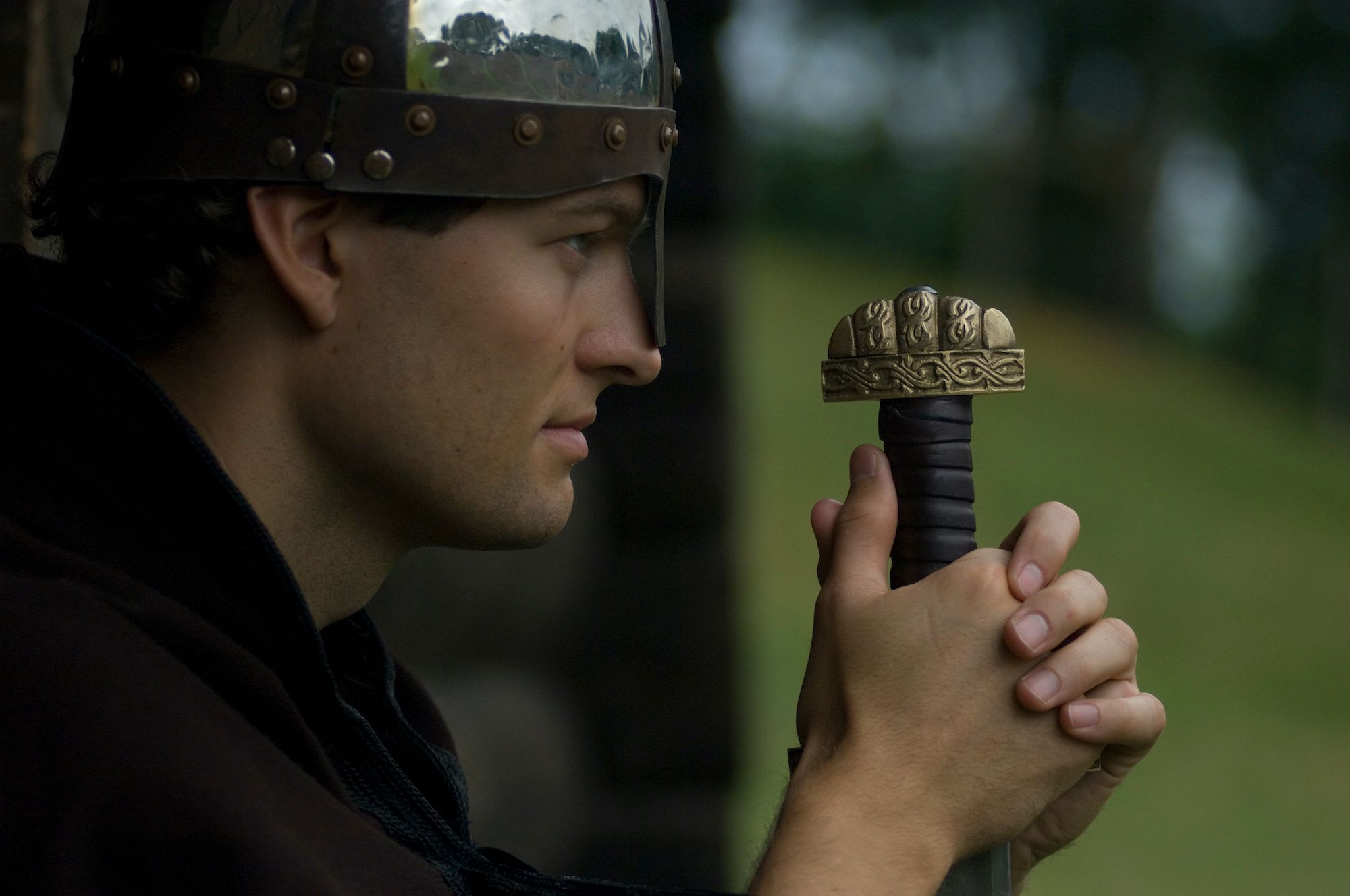 Side profile of a man wearing a metal helmet, holding a sword with intricate engravings.