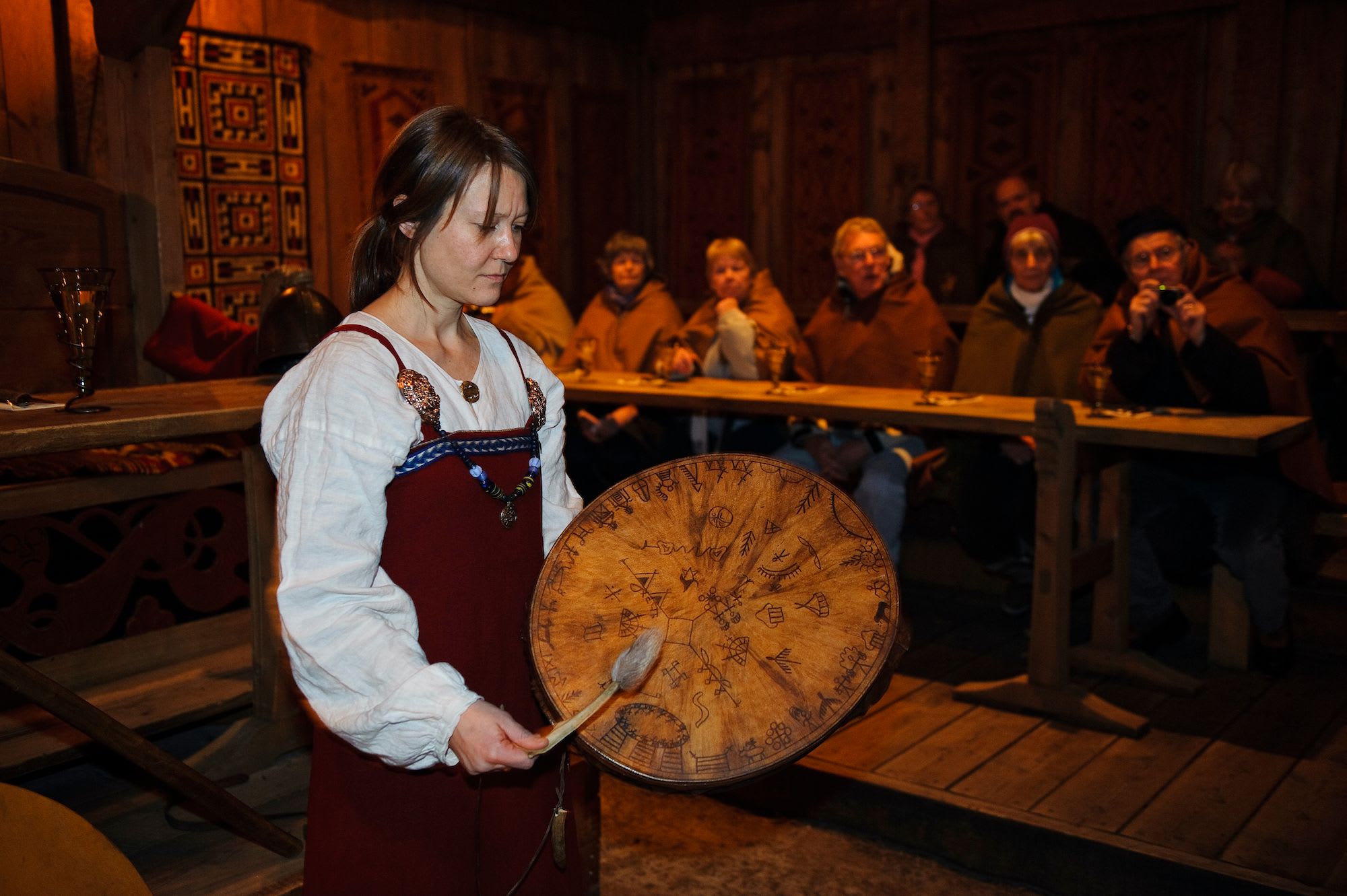 A woman in Viking-era clothing plays a drum with runic symbols in a dimly lit hall, while an audience watches.