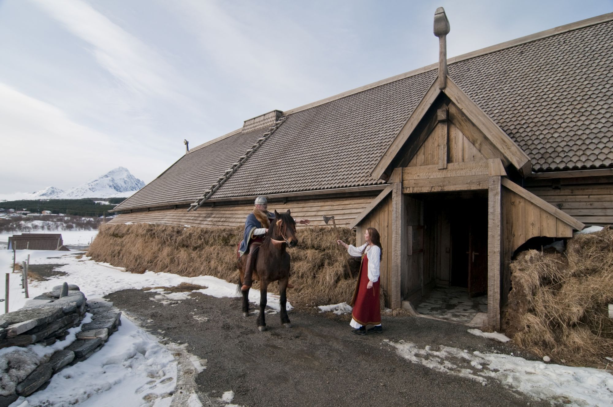 Viking longhouse with wooden walls and sloped roof, man on horse with axe, woman near entrance, snowy mountain in background.