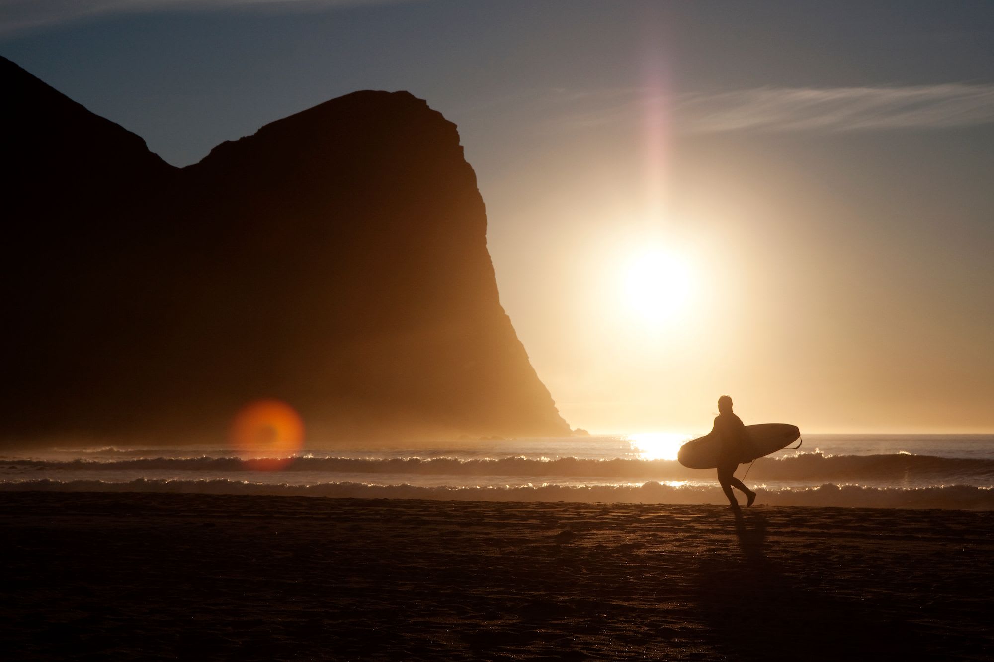 A silhouette of a surfer walking along the beach at sunset, carrying a surfboard.
