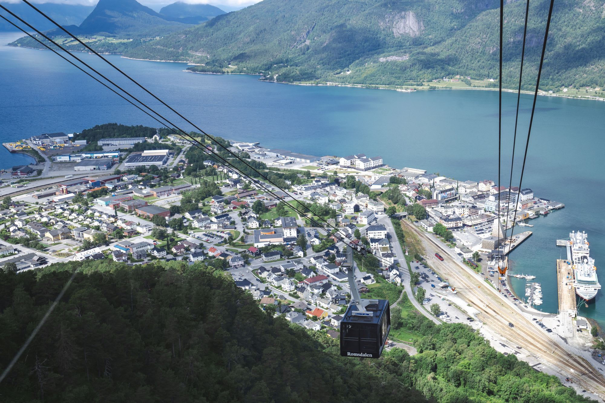 Luftfoto av kystby med taubane over skog og havn omgitt av fjell.