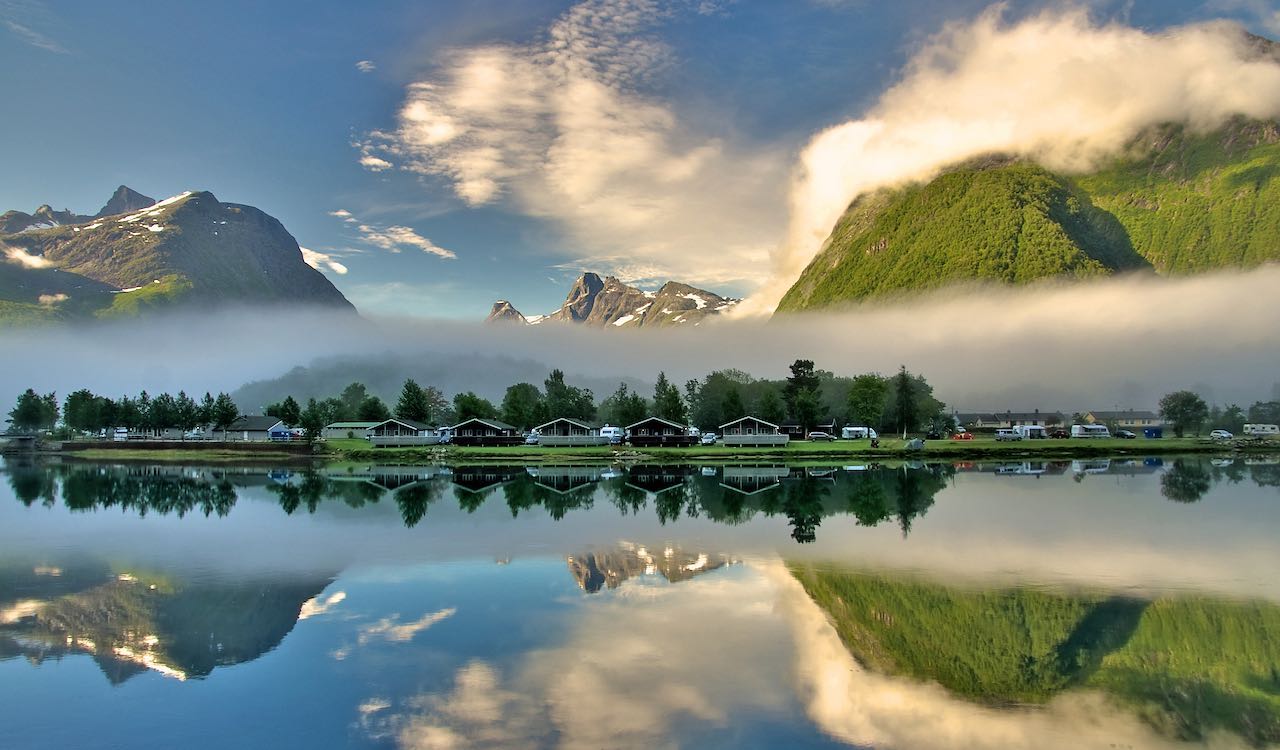 Hytter og campingvogner ved tåkelagt innsjø med speilbilde av fjell.