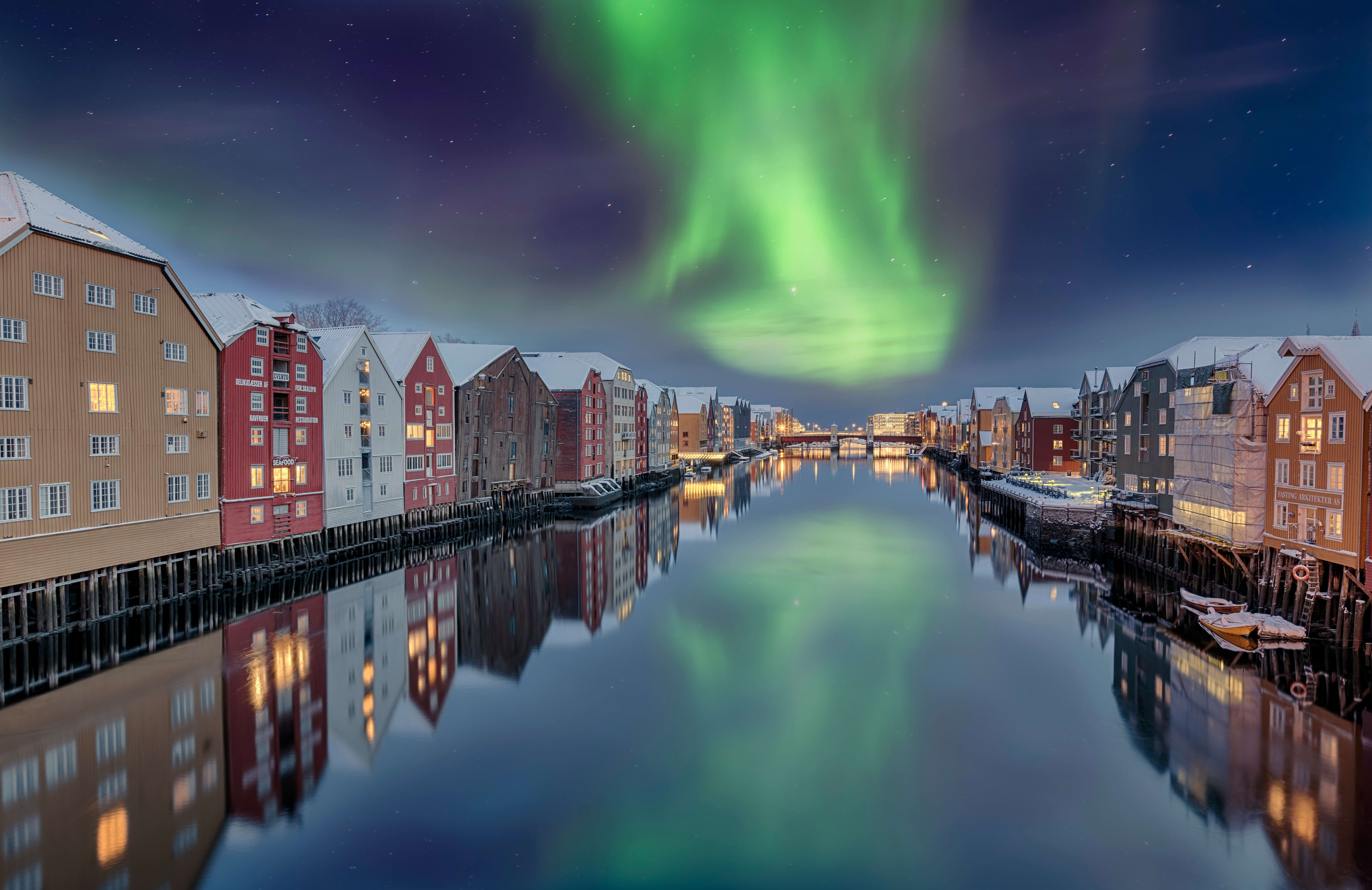 Northern lights reflecting on Nidelva river in Trondheim with colorful wooden buildings along waterfront