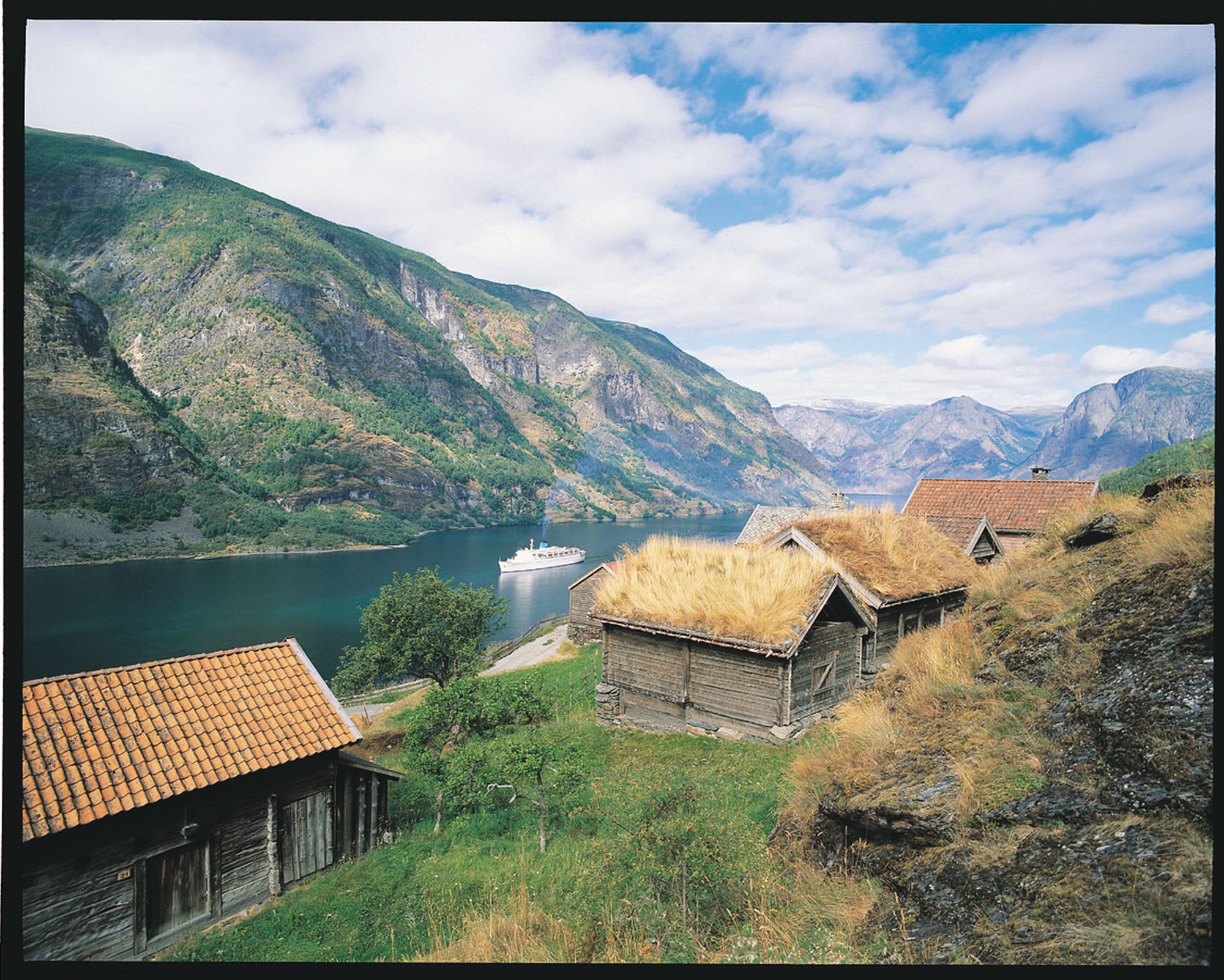 Tradisjonelle trehus med torvtak ved fjorden, med et cruiseskip i det fjerne og fjell i bakgrunnen.