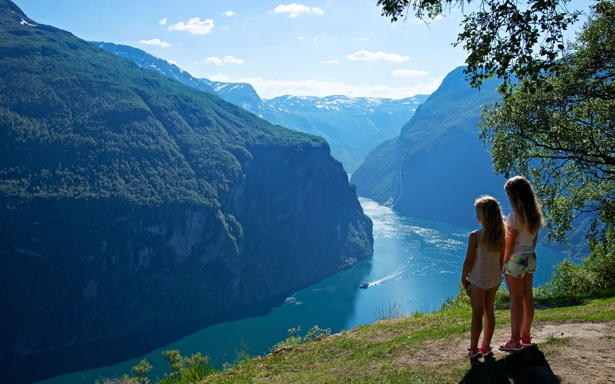 To jenter ser på norsk fjord, klipper dekket med grønt, rolig blått vann som reflekterer den lyse himmelen.