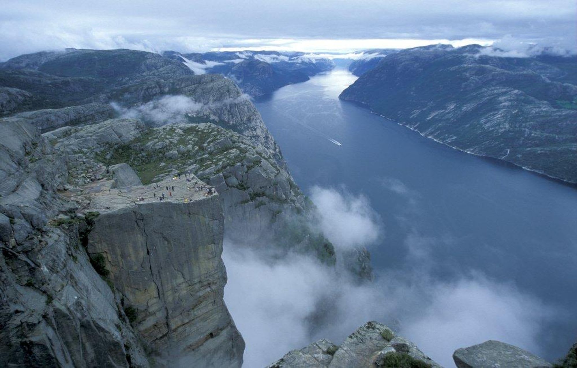Luftbilde av Preikestolen (Prekestolen), med folk som står på klippe med fjord i bakgrunnen, tåkete fjell og fjord.