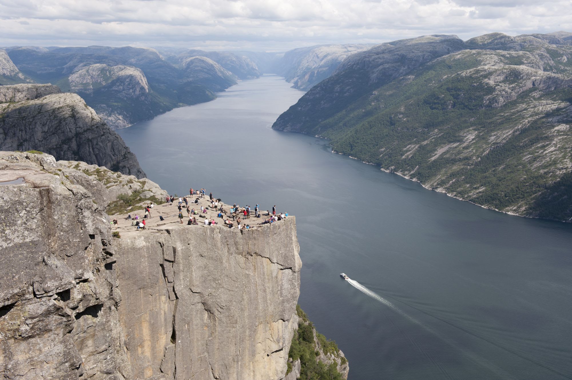 Aerial view of popular Norwegian hiking destination, people standing on cliff with fjord below, small boat sailing through.