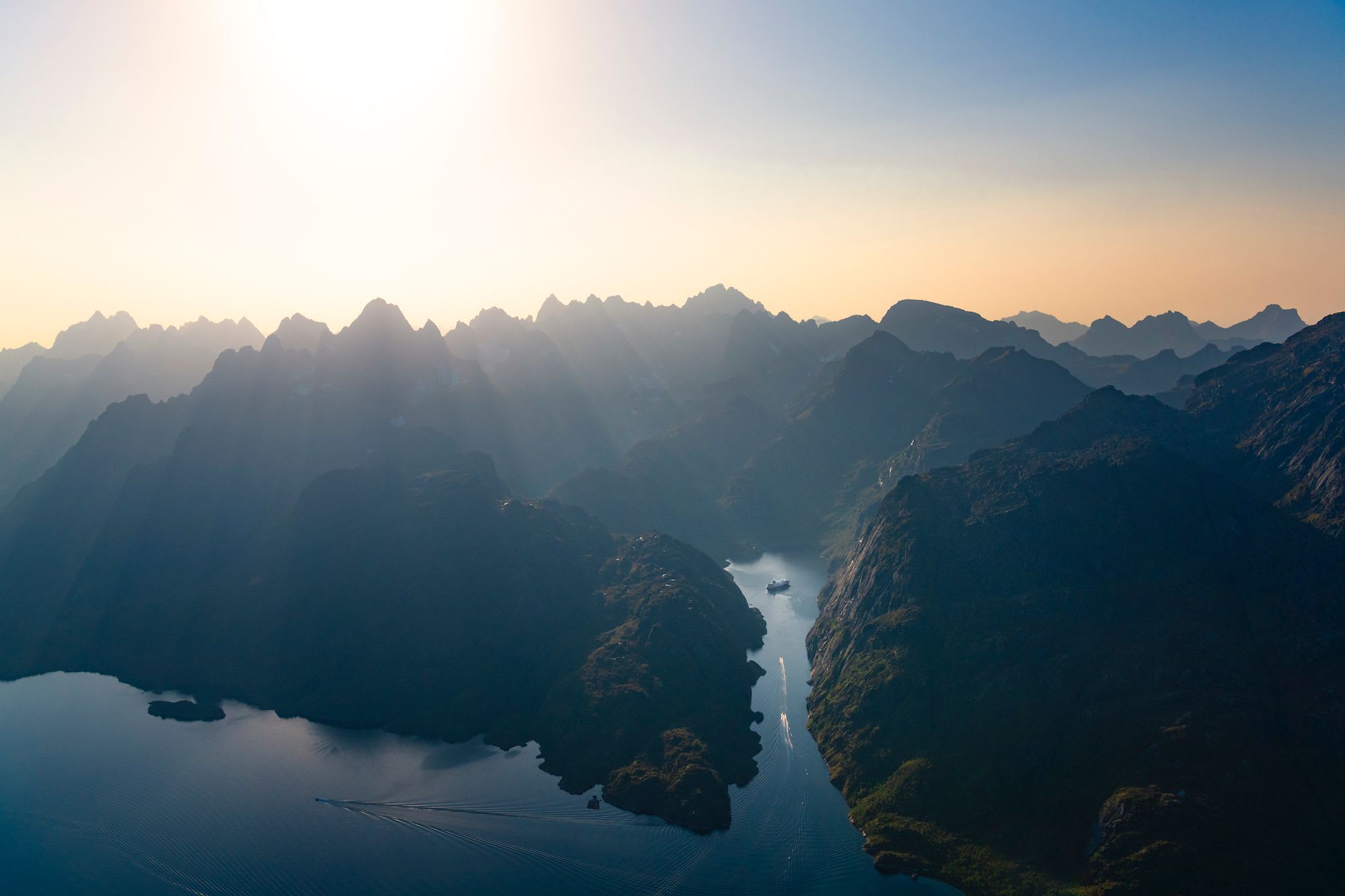 Aerial view of rugged mountains at sunrise with sun rays piercing peaks, fjord, and a ship sailing through calm water.