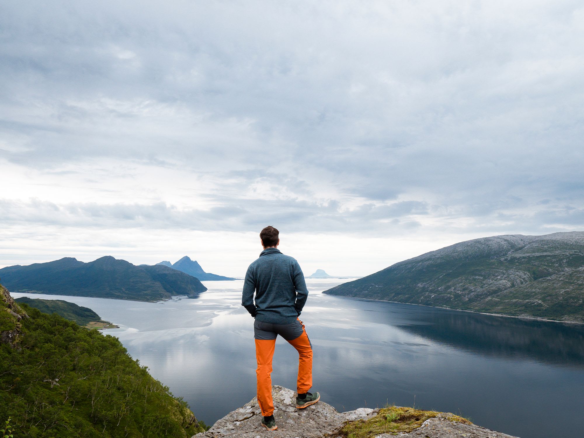 Person på steinete klippe som ser ut over fjord med grønne bakker og fjerne fjell under en skyet himmel, som gir en følelse av eventyr.