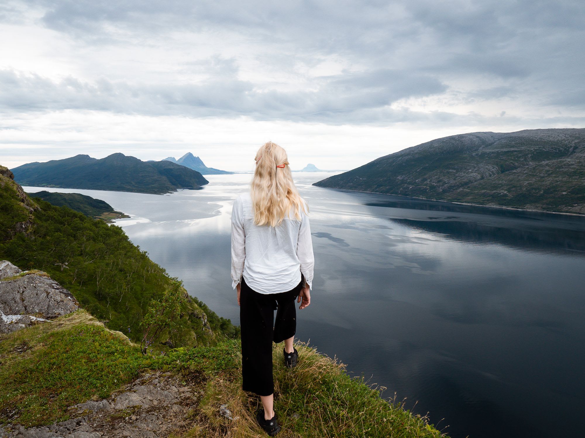 A woman standing on the edge of a cliff, gazing at a fjord with dramatic mountains and calm waters.
