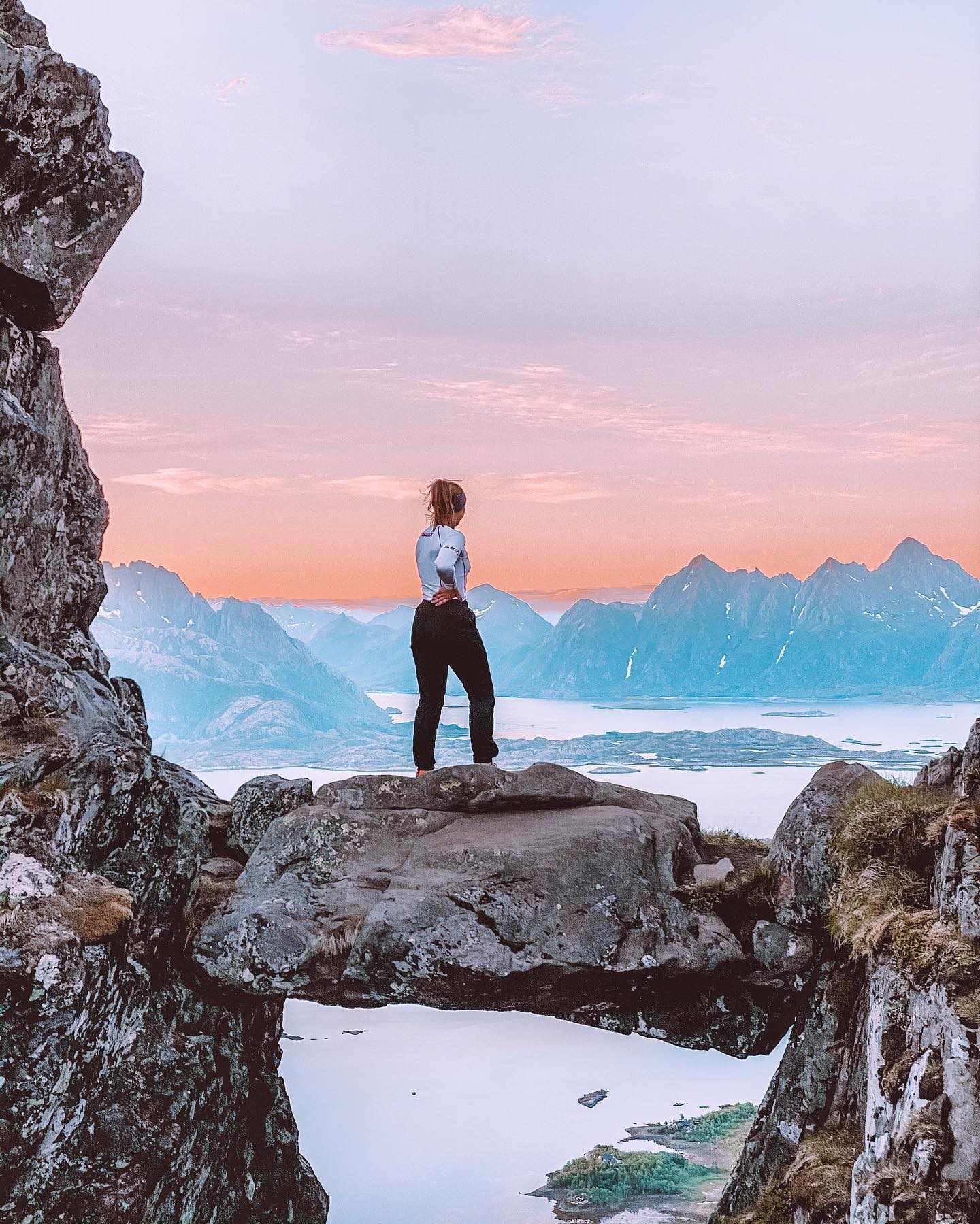 En kvinne står på en naturlig steinbro og ser utover en fantastisk fjord- og fjellutsikt ved solnedgang.