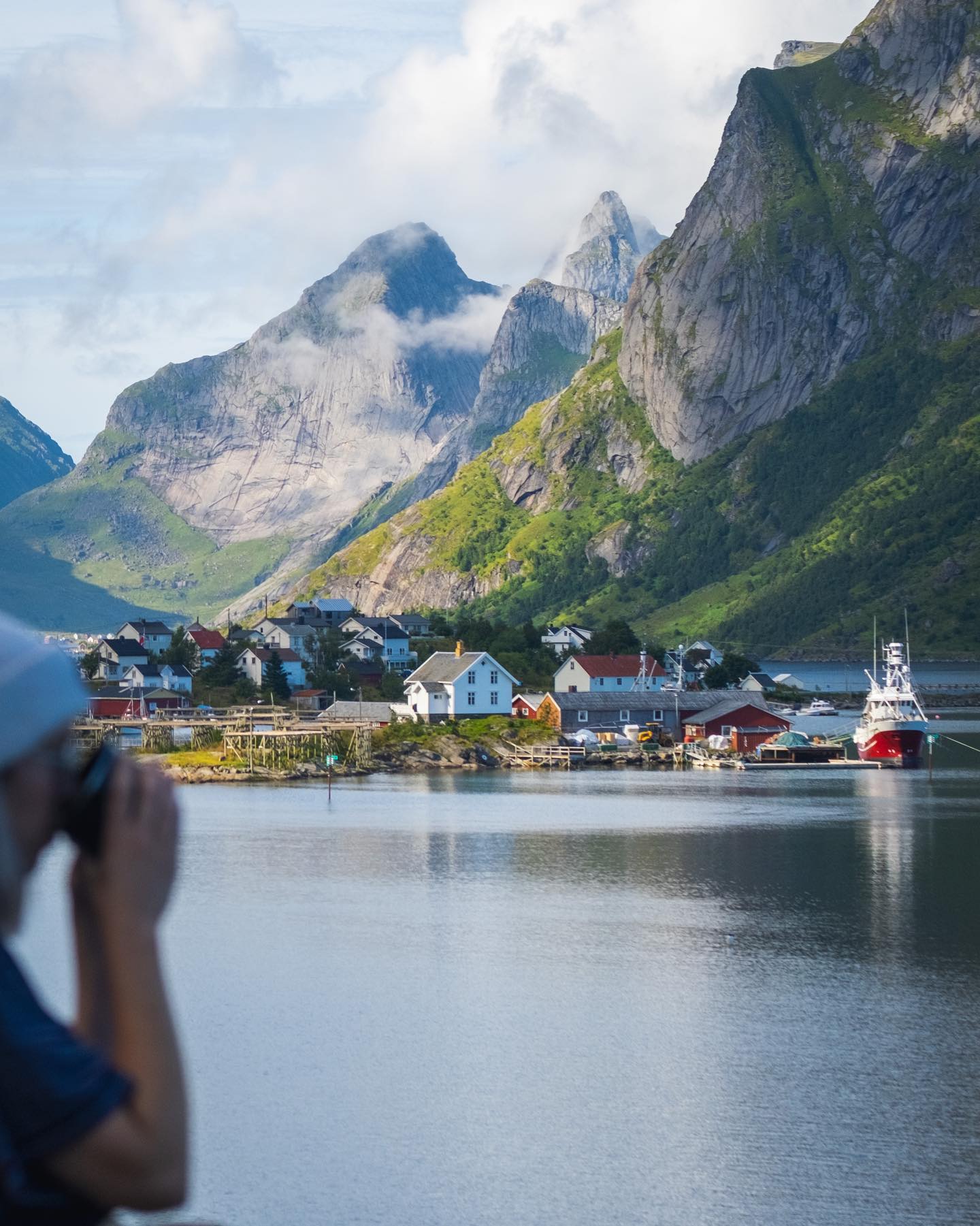 Person fotografiert Dorf am Fjord mit grünen Bergen und Wolken.