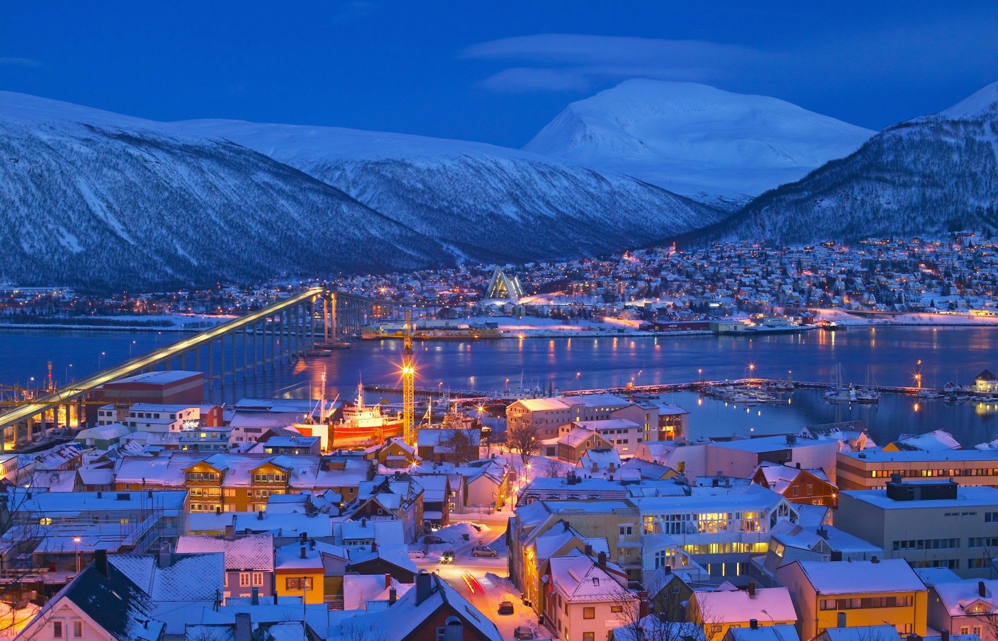 Snowy city at dusk with lit bridge and colorful houses.