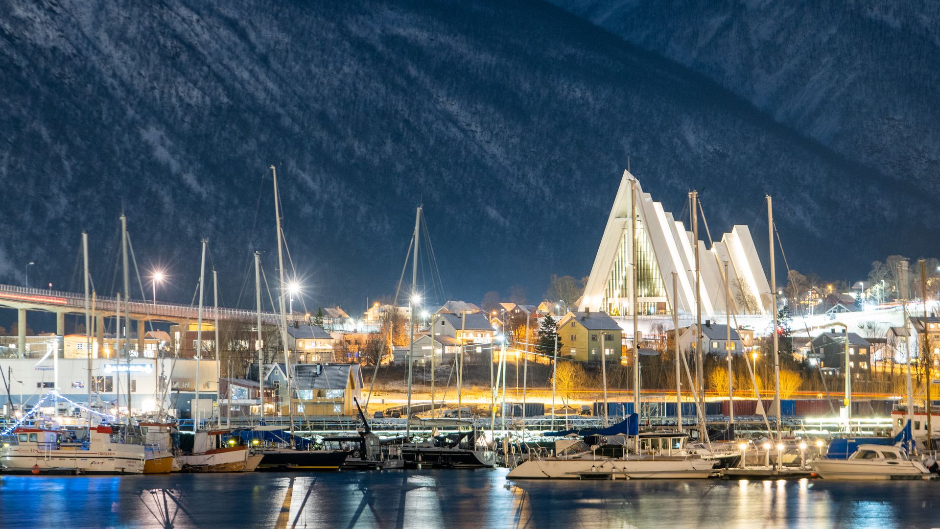 Die beleuchtete Eismeerkathedrale in Tromsø bei Nacht, mit Fischerbooten im Hafen darunter und schneebedeckten Bergen, die sich im Hintergrund erheben.