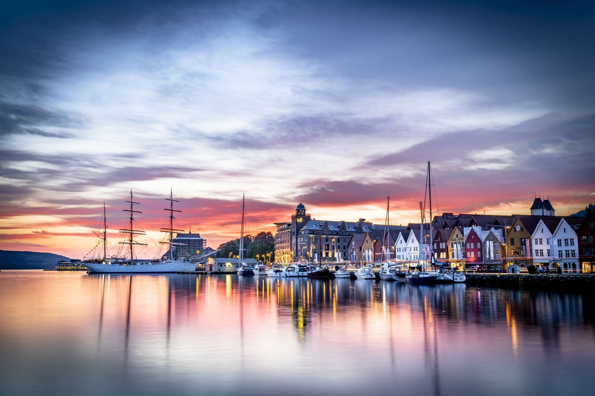 Sunset harbor scene with colorful wooden buildings, sailboats docked, and their reflections in calm water under a dramatic sky.
