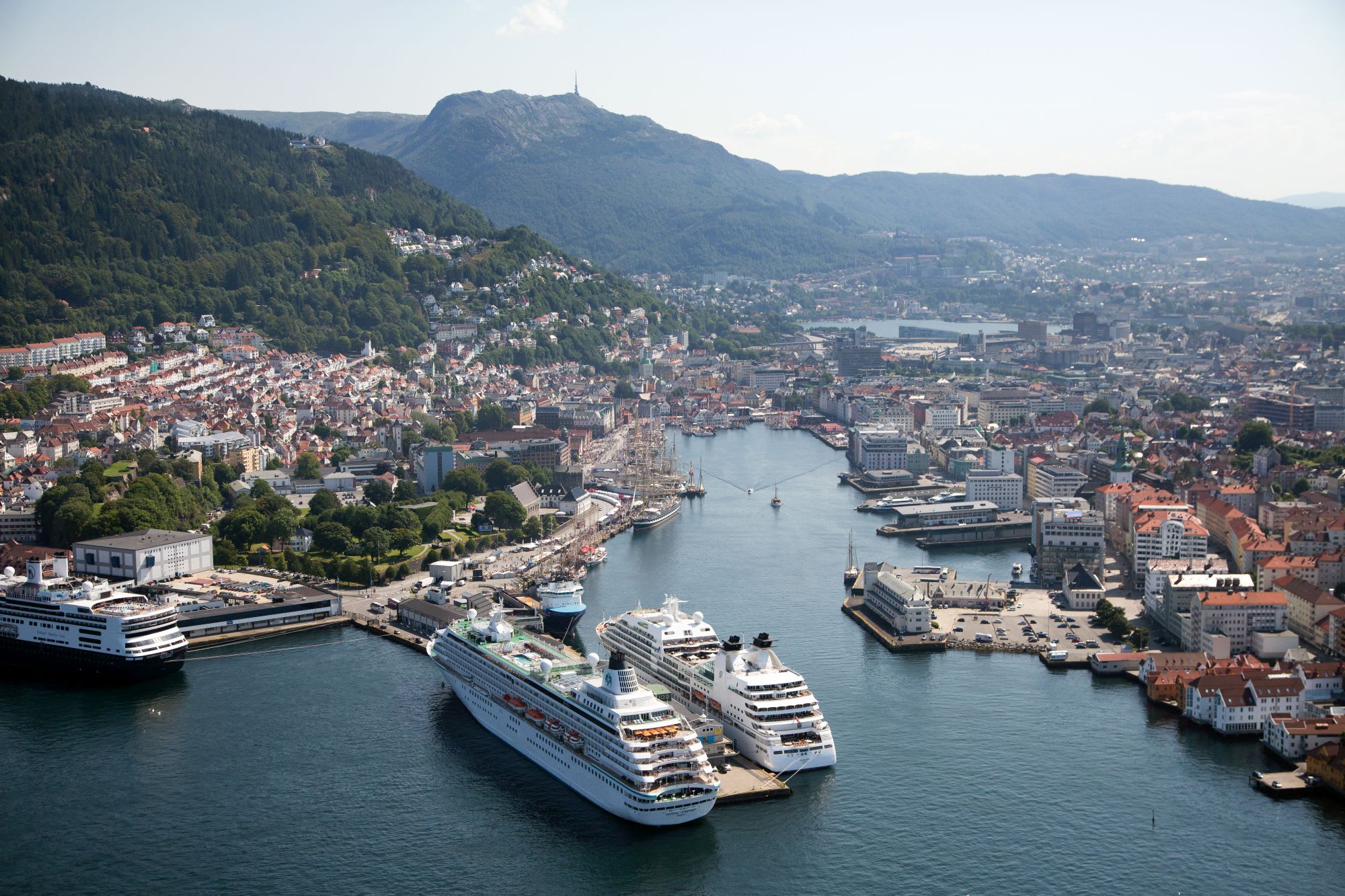 Aerial view of a bustling coastal city with cruise ships docked in a lively harbor, colorful buildings, and green hills under a sunny sky.