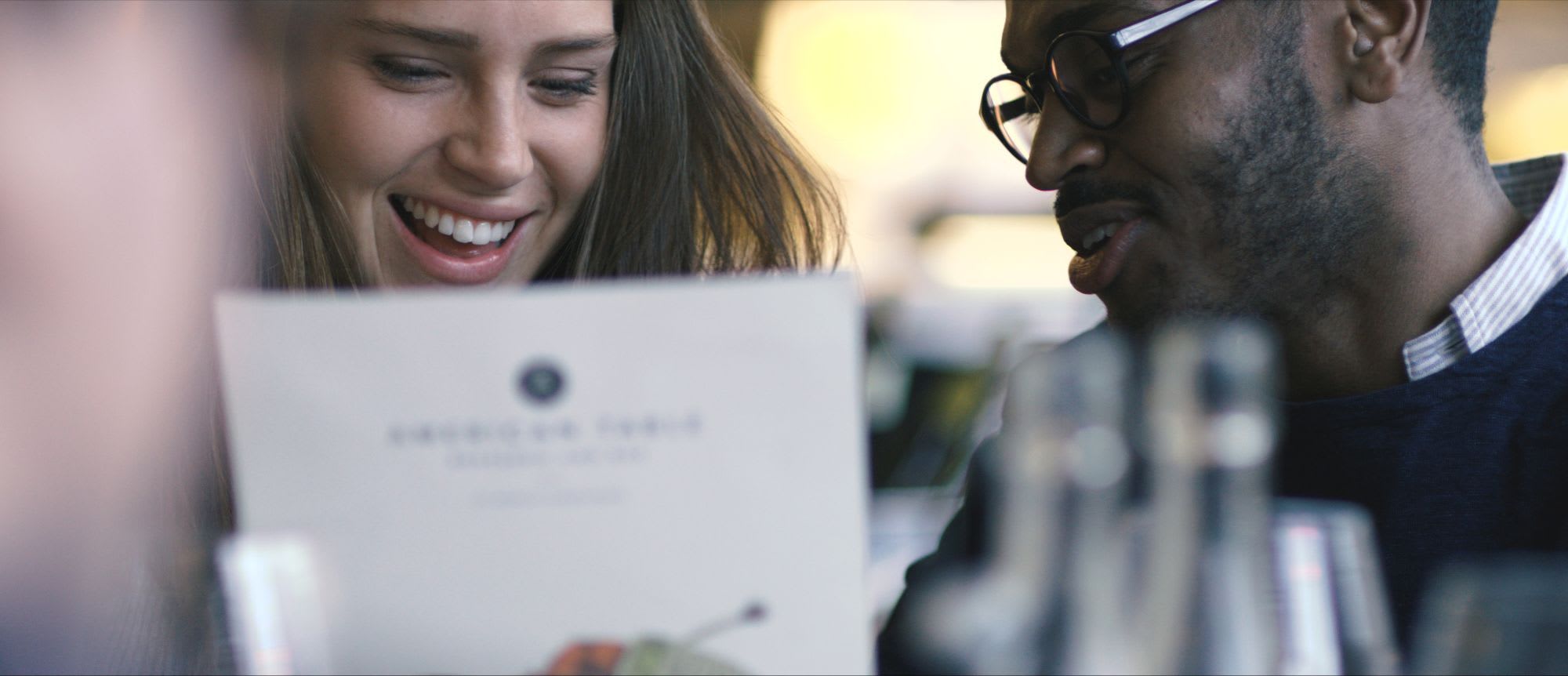 Two people smiling while looking at a menu in a cozy, relaxed atmosphere with blurred glassware and warm indoor lighting.