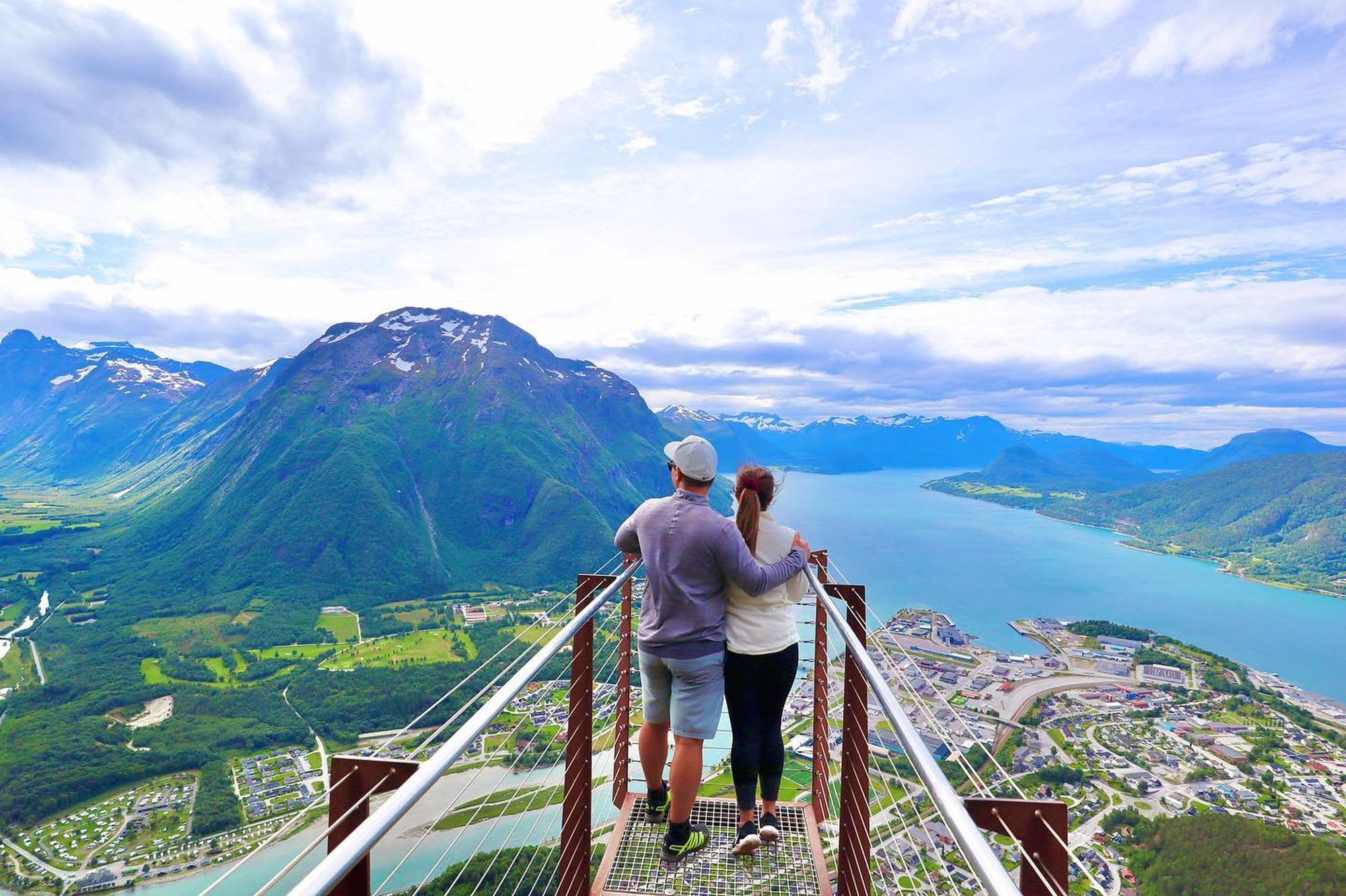 Couple standing on a viewing platform, embracing while looking at a fjord with green mountains, snow patches, and a small town below.