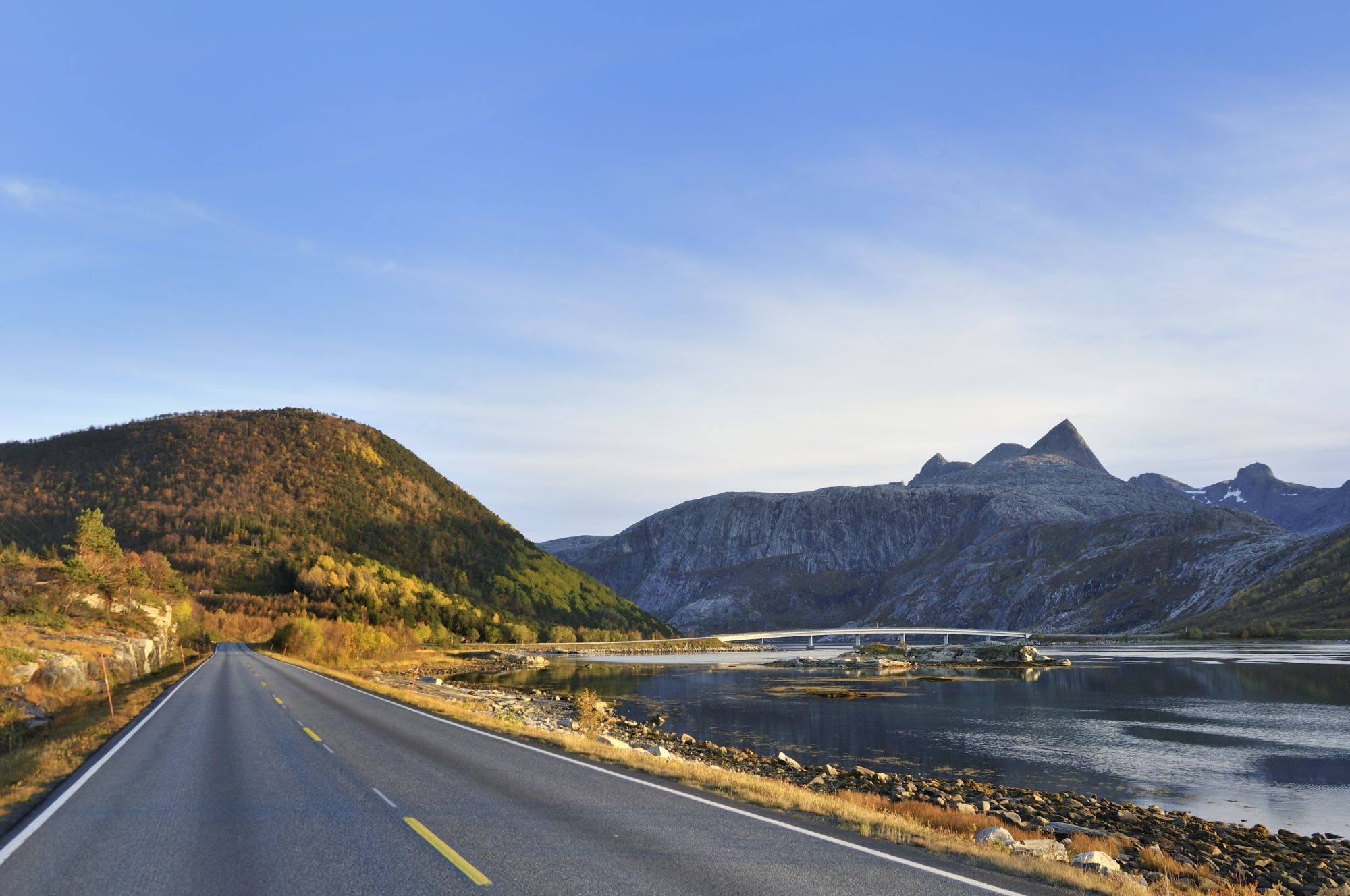 Open road leading to a white bridge over calm water, surrounded by green hills and rocky mountains under a clear, blue sky.