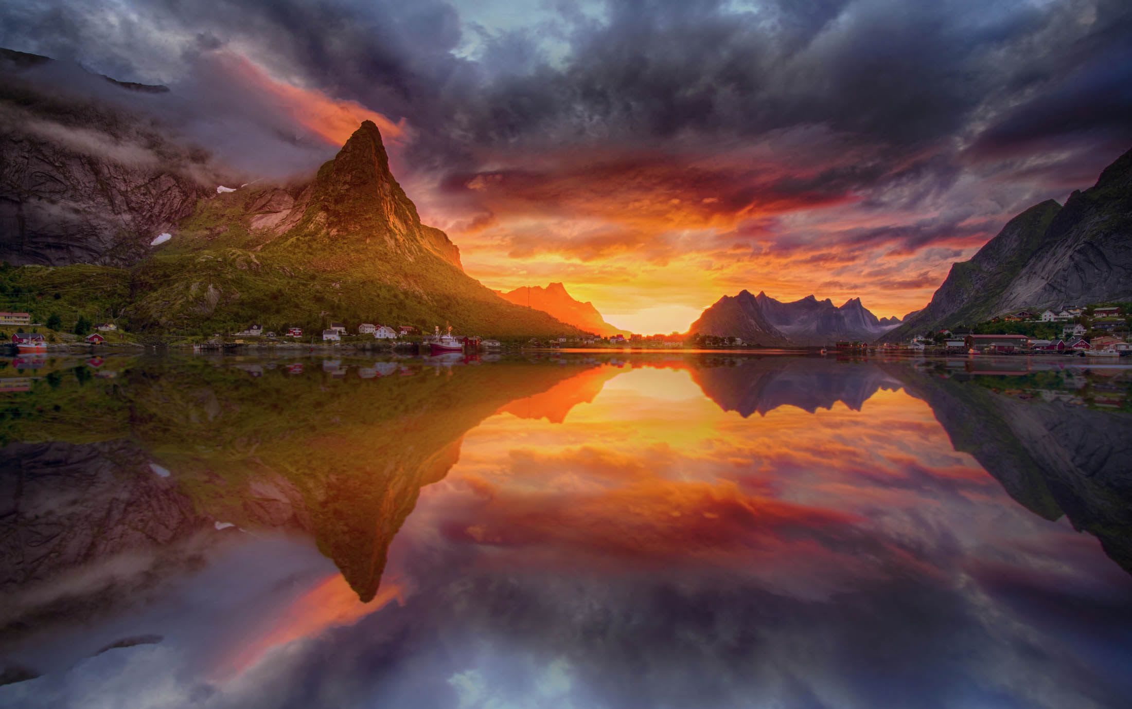 A stunning sunset over a serene fjord, with mountains surrounding a small village along the shoreline.