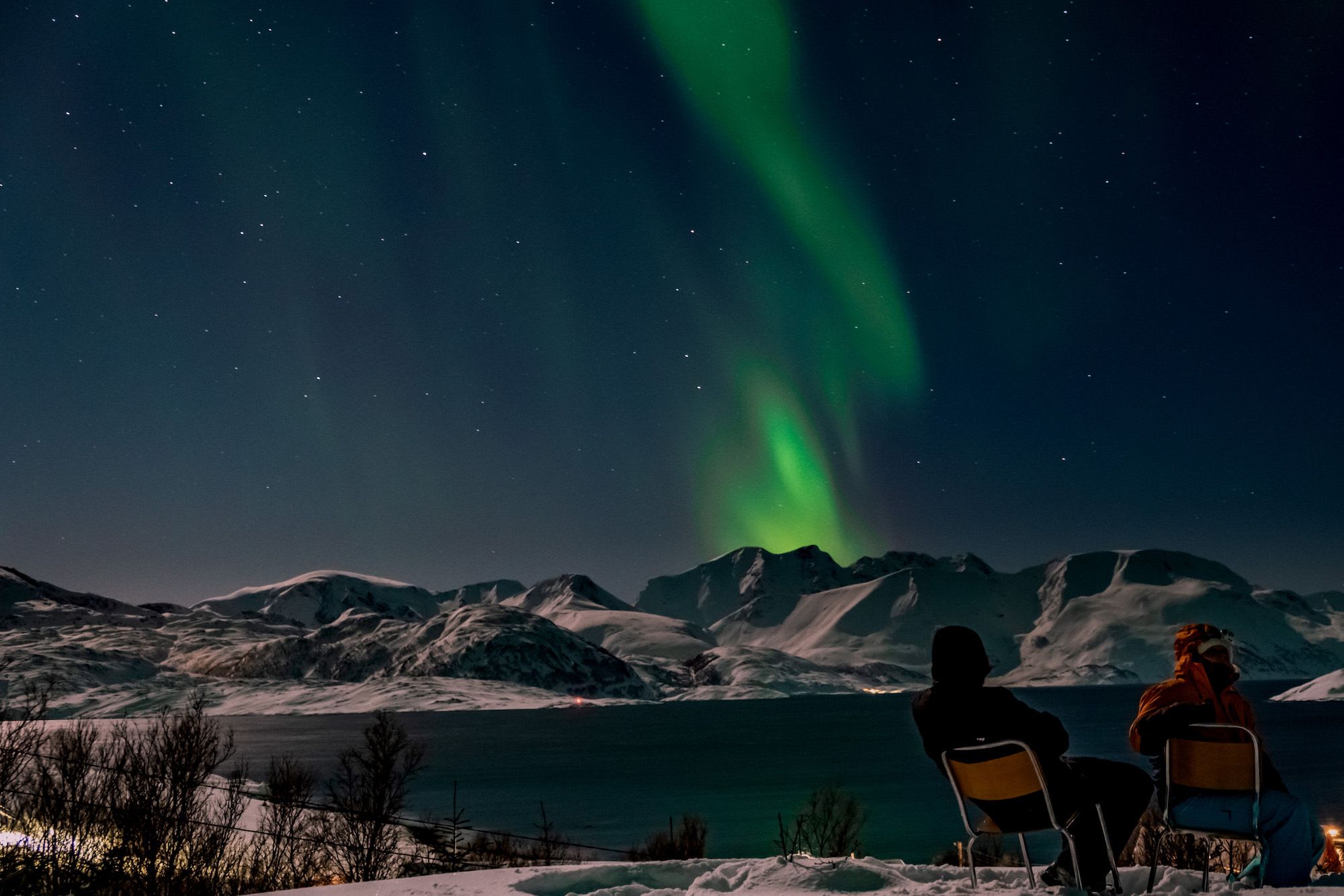 To personer sitter i snøen og ser på nordlyset.