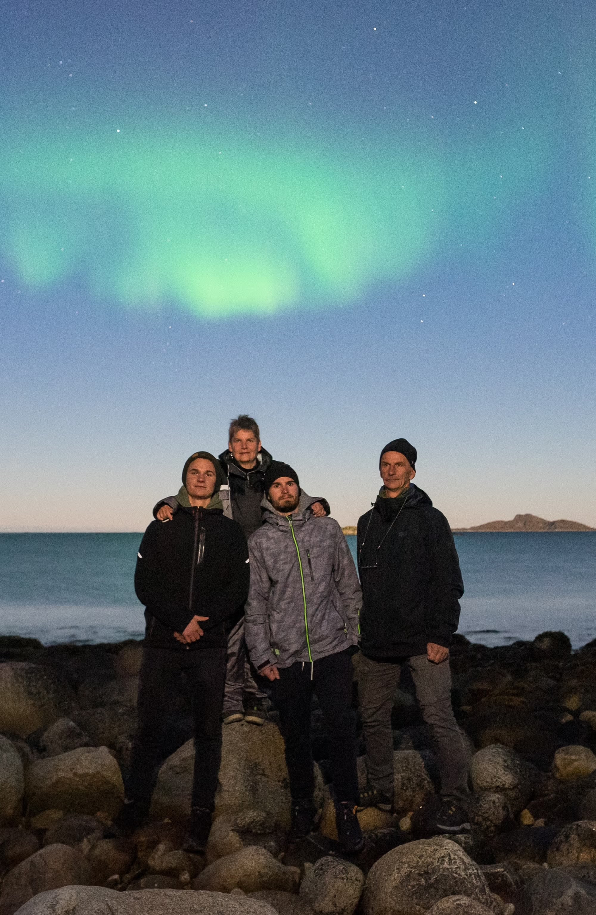 Fire familiemedlemmer på steinete kyst med grønne nordlys som speiler seg i vannet