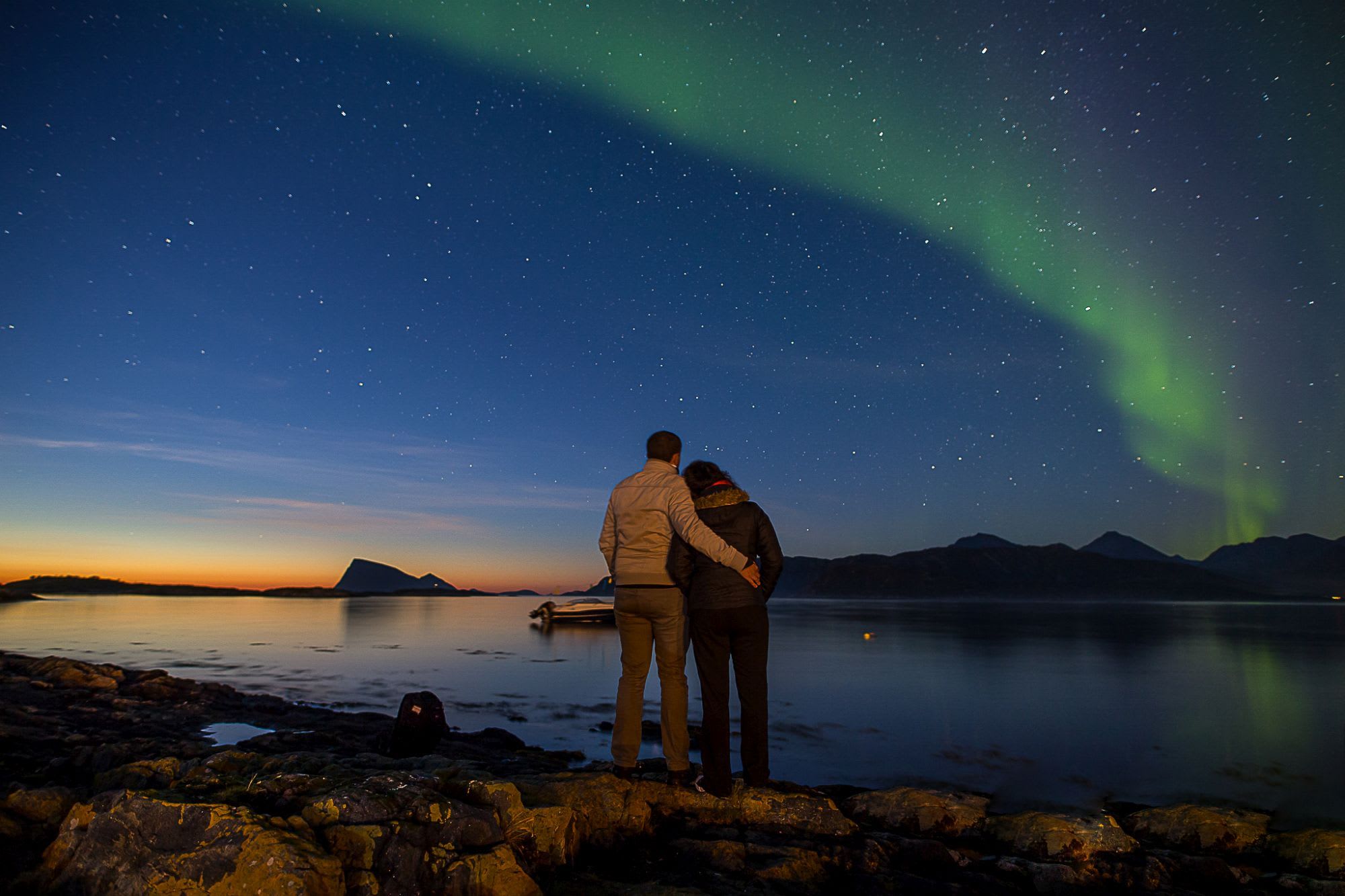 Et par omfavner hverandre under nordlyset på en steinete strand. Aurora speiler seg i vannet, med fjell i bakgrunnen.