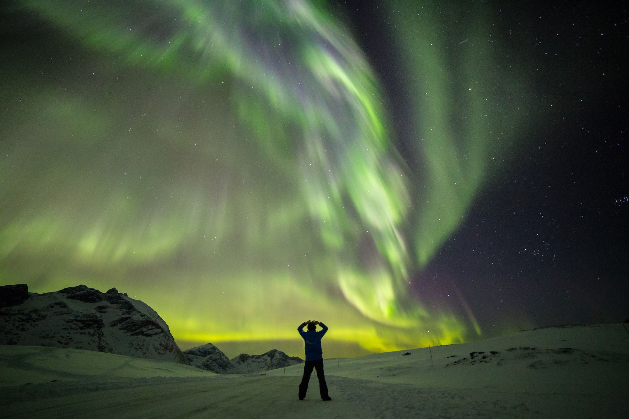 Person standing in snow with arms raised under bright green and yellow Northern Lights, surrounded by snow-capped mountains.