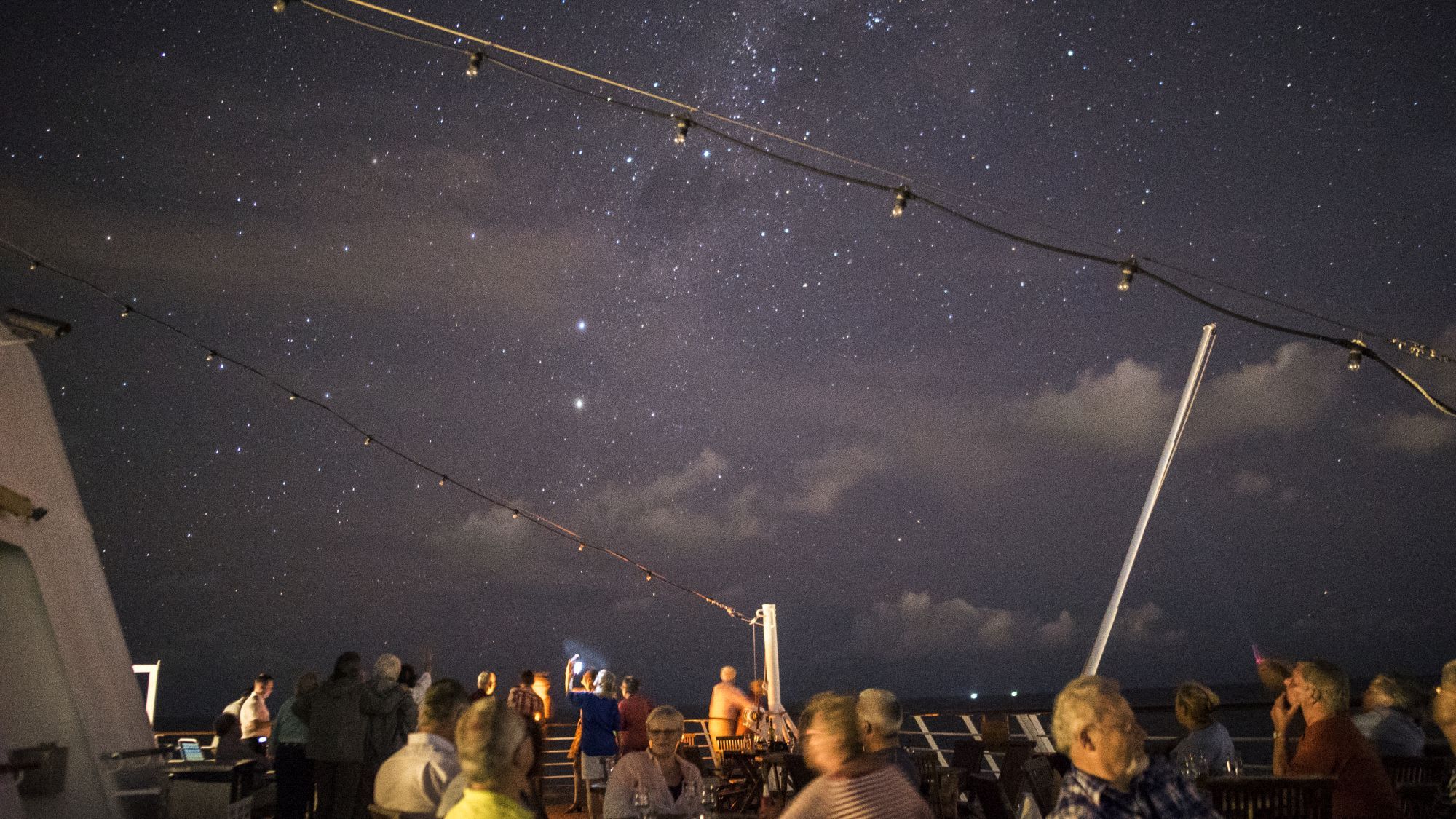 Gruppe samlet på skipets dekk om natten, noen sitter og ser på stjernehimmelen med lyslenker over hodet.