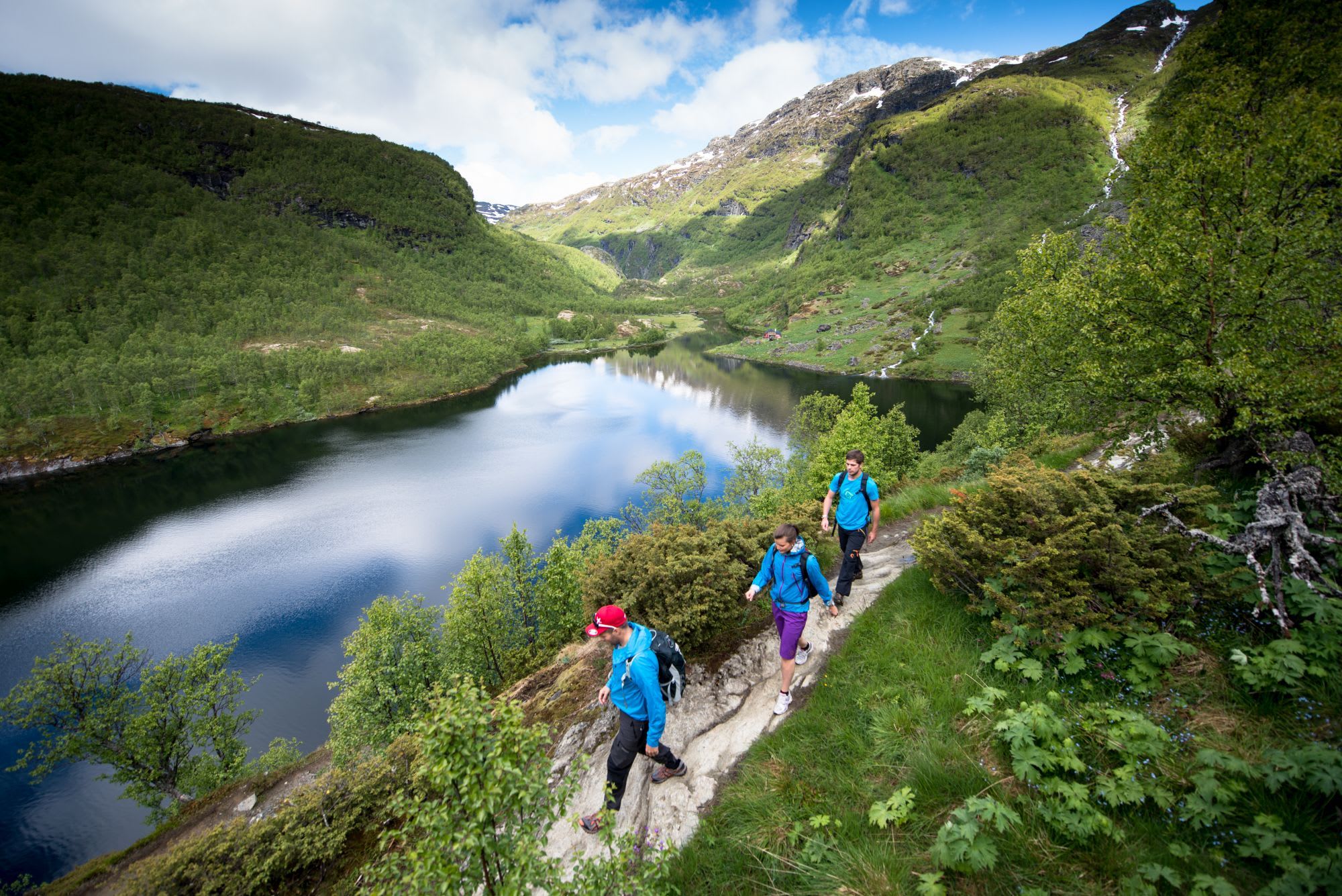 Hikers in colorful gear on a trail by a lake in a lush valley framed by hills and snow-capped peaks.