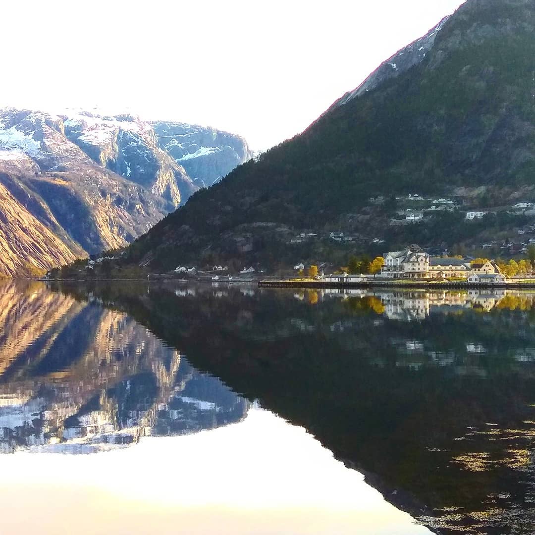 Snødekte fjell og hotell speiles i stille fjordvann.