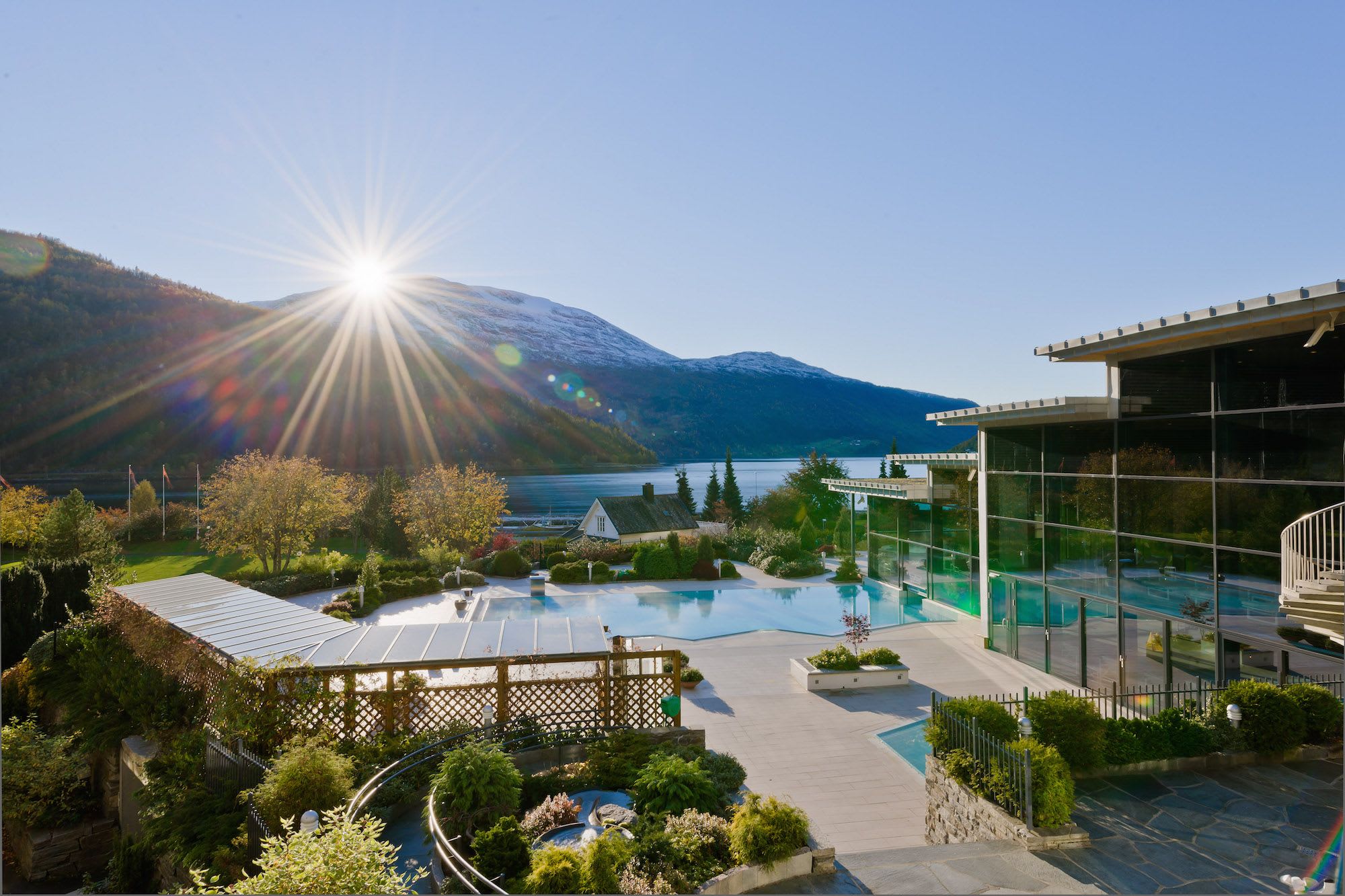 Moderne glassveggsresort med utsikt over turkis fjord og snøkledde fjell ved soloppgang/solnedgang.