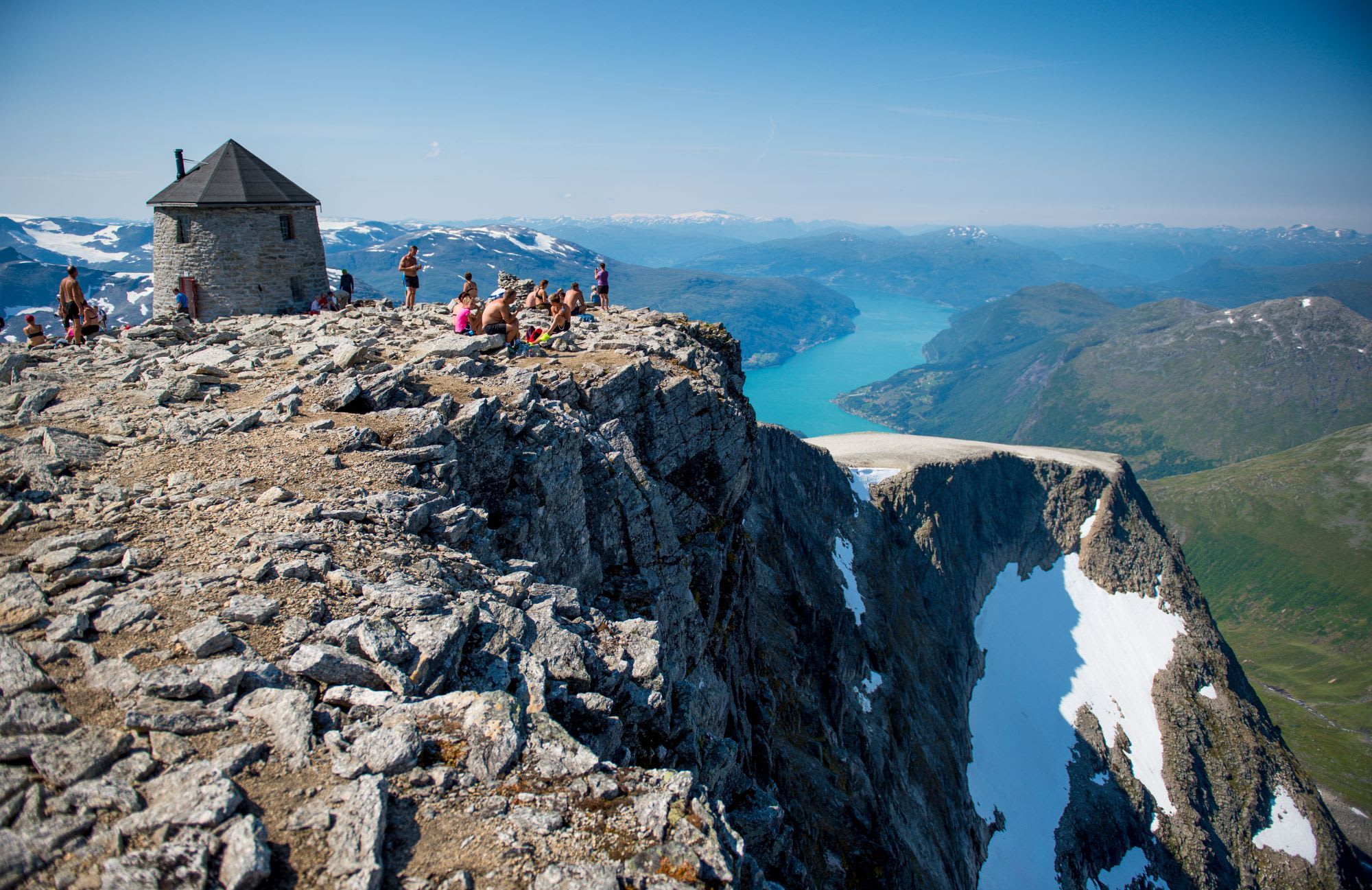 Mountain summit with people sunbathing, stone cabin, and turquoise fjord framed by snow-capped peaks.