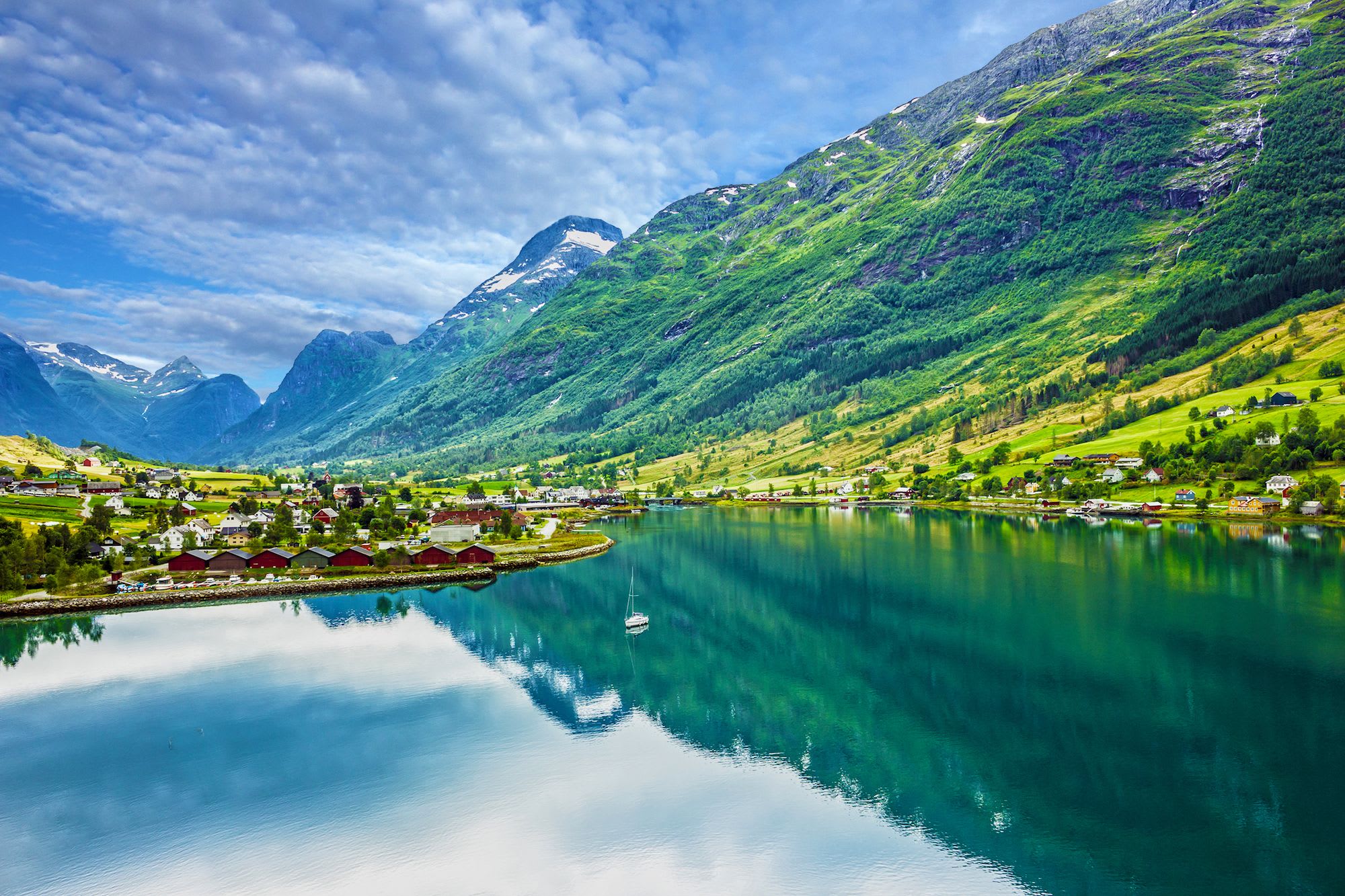 Calm fjord village with boat, green hills, and snow-capped peaks mirrored in water.