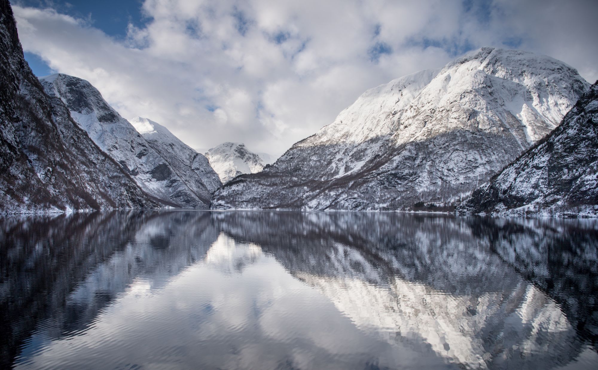Calm fjord reflecting snow-capped mountains and cloudy sky, creating a mirror effect.