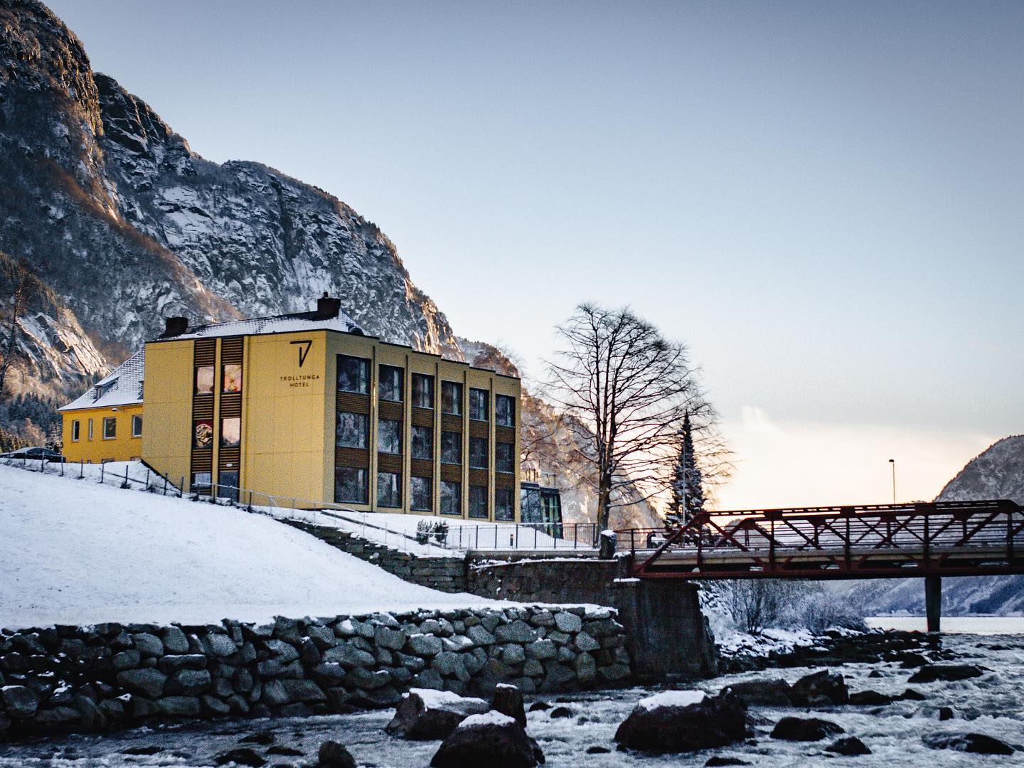 Gult Trolltunga Hotel ved elv og rød bro i snødekt landskap.
