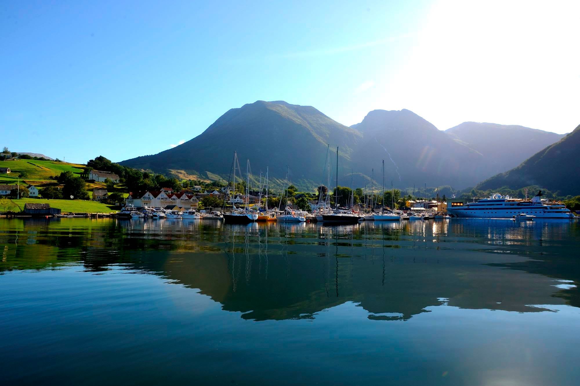 Peaceful marina with sailboats, red-roofed village, and green mountains reflected in calm water.
