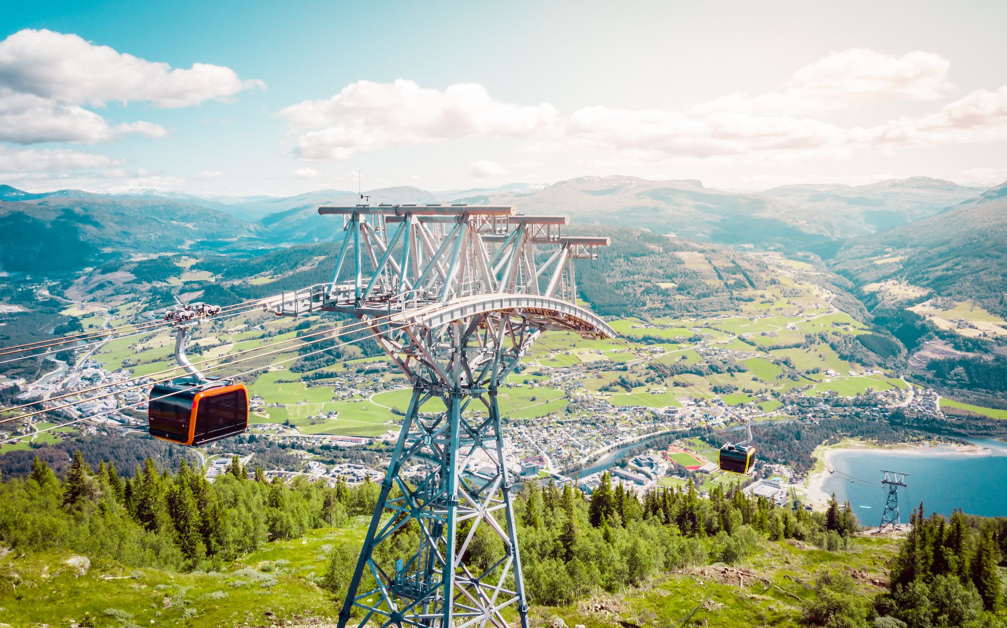 Red gondolas travel above green valley and lake with mountains beyond.